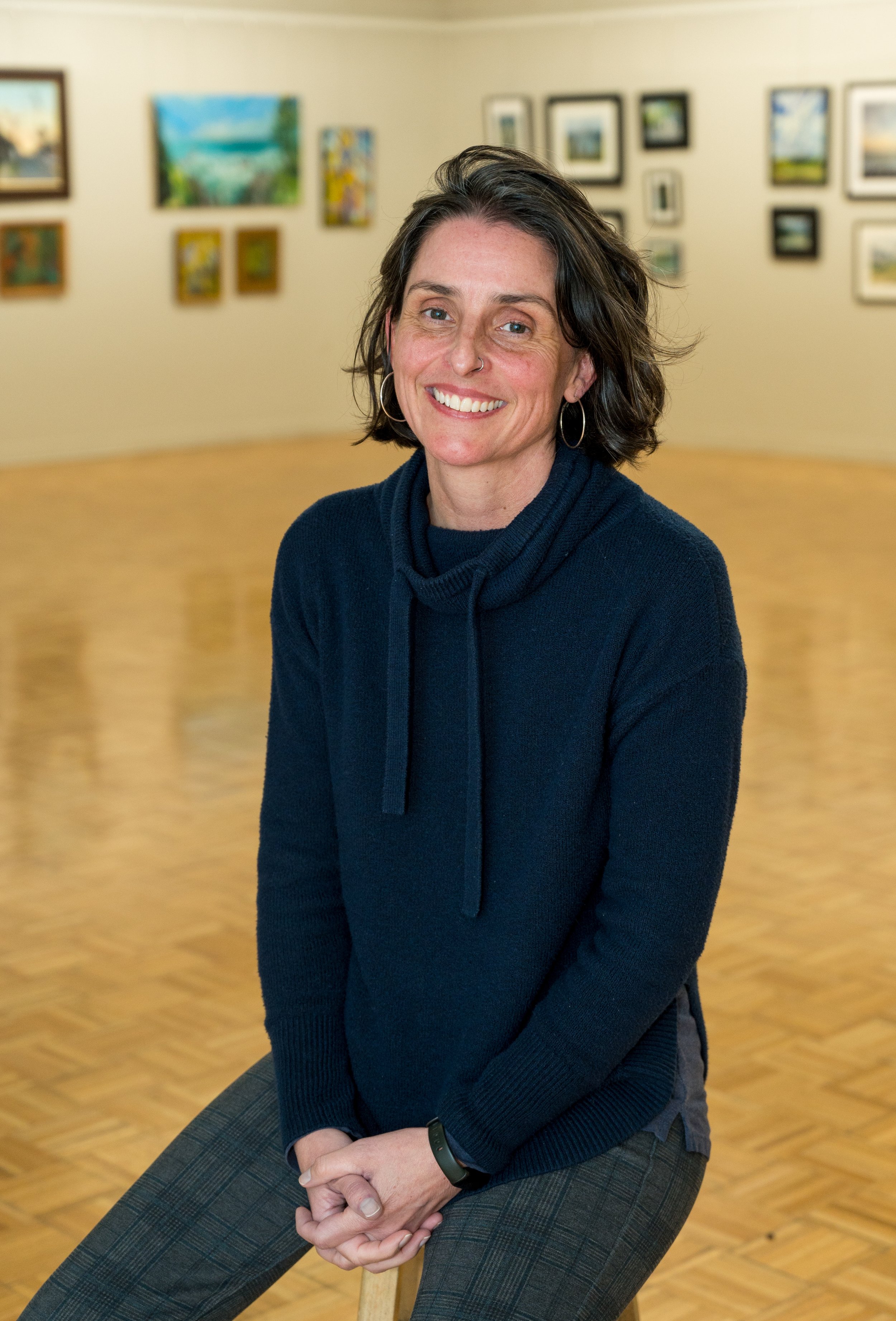 A woman with curly brown hair and hoop earrings sitting on a stool in an art gallery, wearing a navy blue sweater and plaid pants, smiling at the camera.