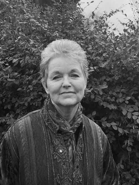 Older woman with short hair wearing a patterned jacket and scarf, standing in front of dense bushes.