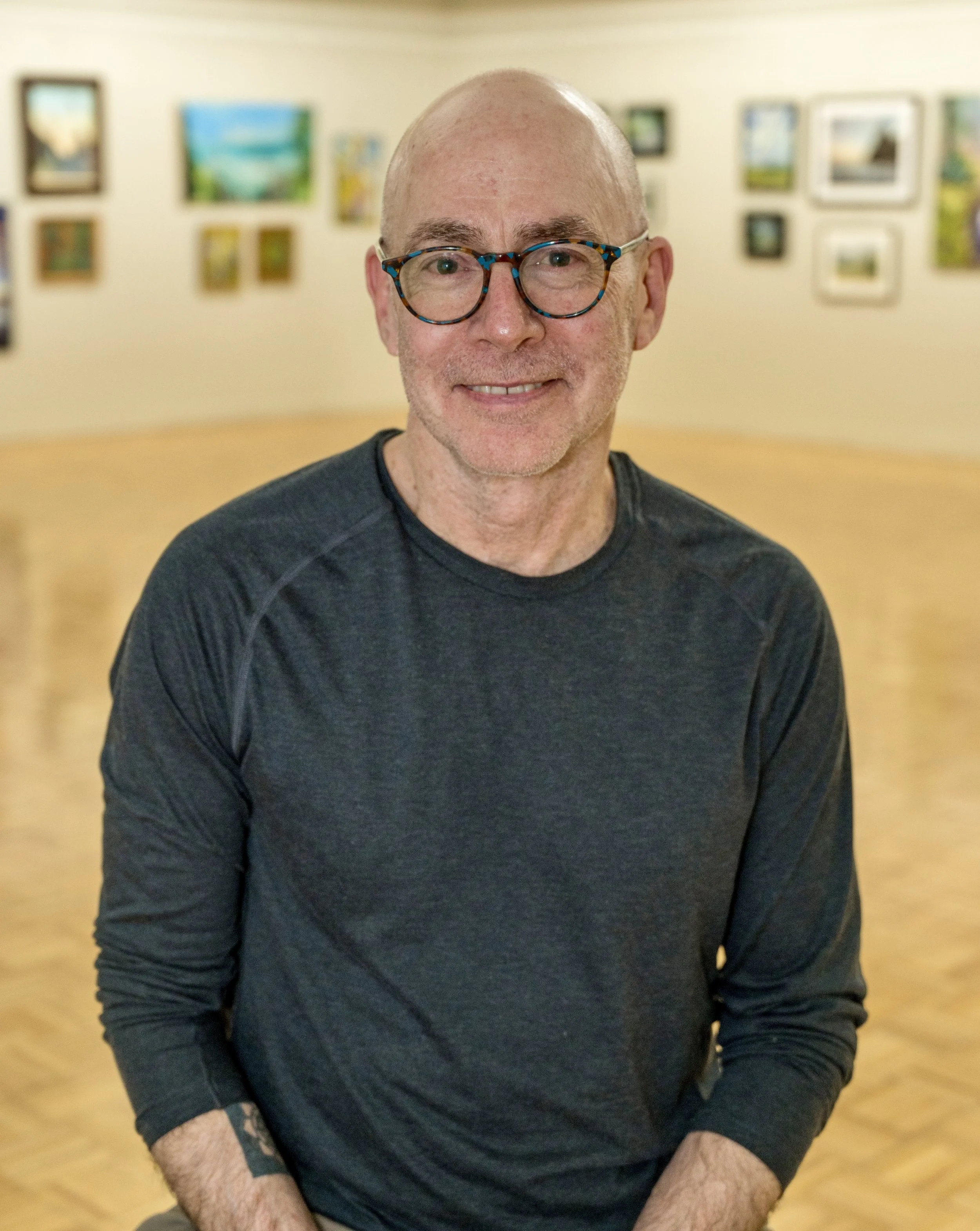 A smiling man with glasses and a black long-sleeve shirt, sitting in an art gallery with colorful paintings on the wall behind him.