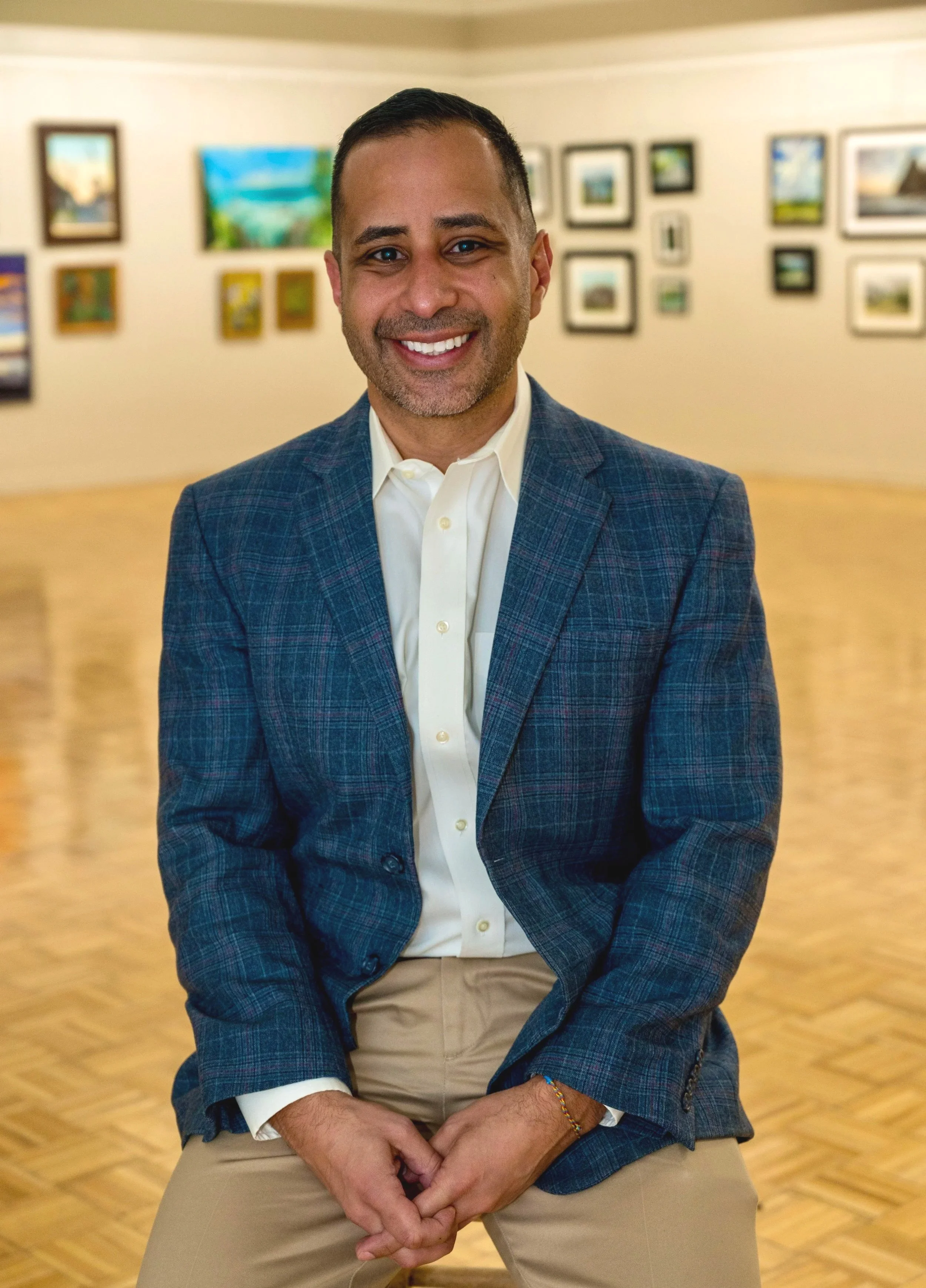 A man in a blue plaid blazer and beige pants, sitting in an art gallery with framed paintings on the wall behind him, smiling at the camera.