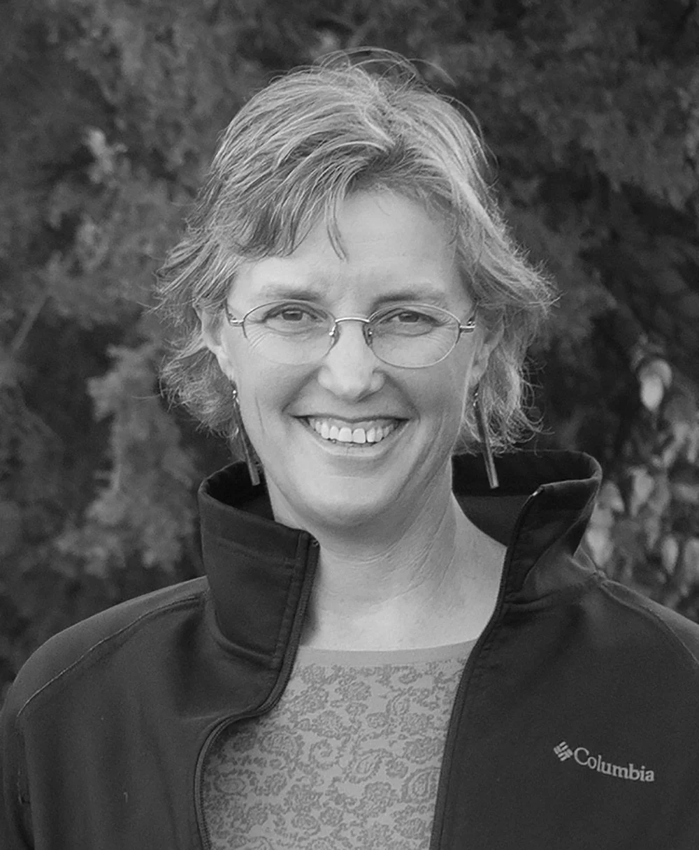 Black and white photo of a woman with short, wavy hair, glasses, and earrings, smiling outdoors, wearing a Columbia jacket and patterned shirt.