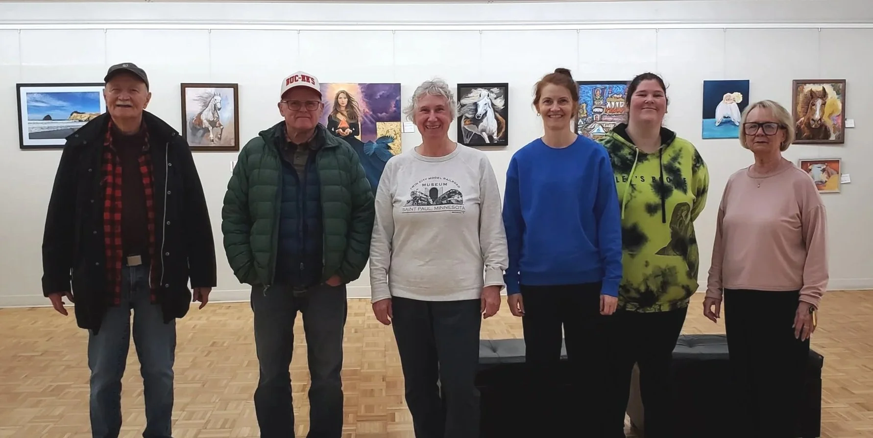 Six people standing side by side in an art gallery with colorful paintings on the wall behind them.