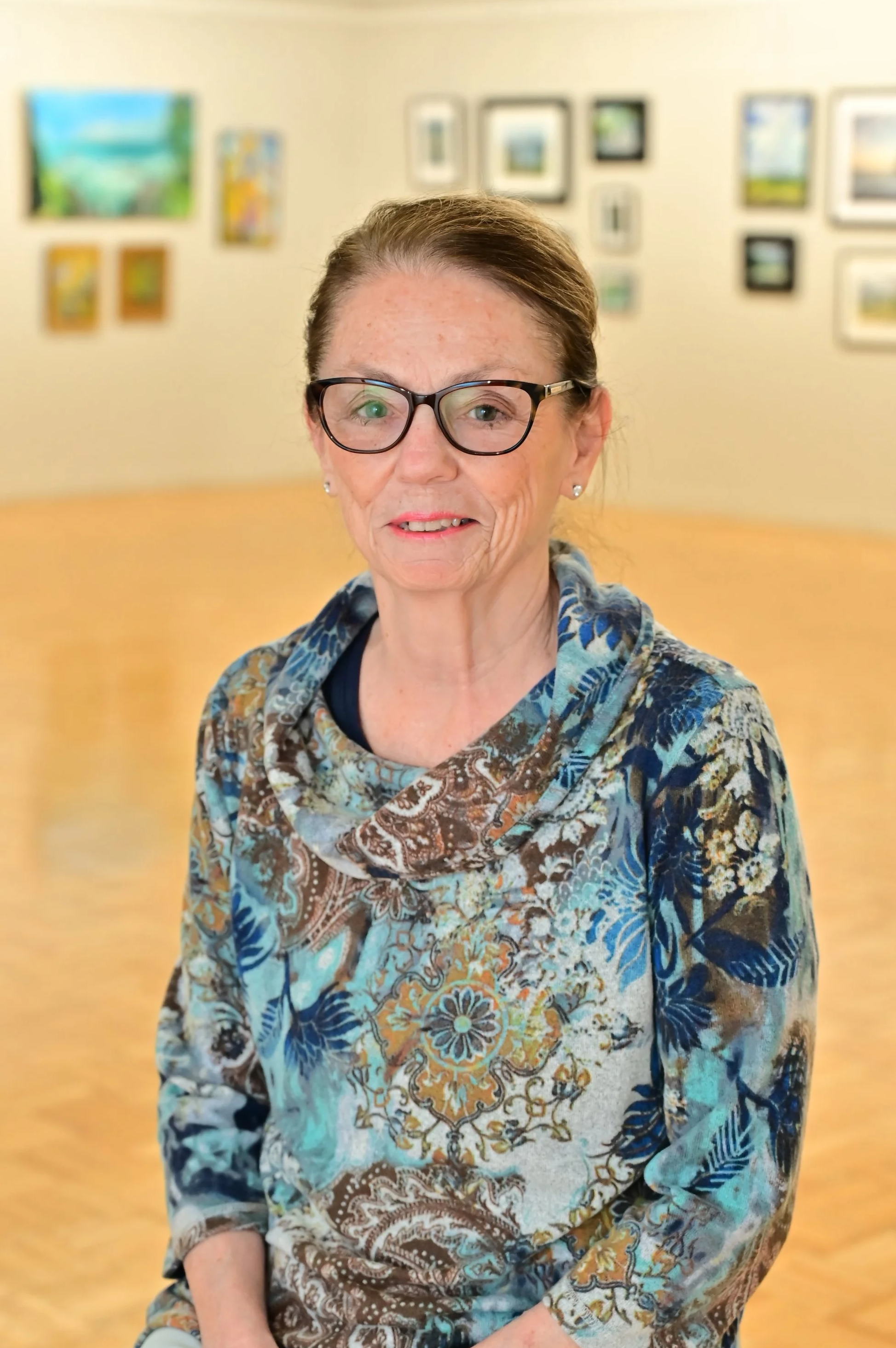 A woman with glasses and a colorful patterned blouse sitting in an art gallery with framed paintings on the wall behind her.