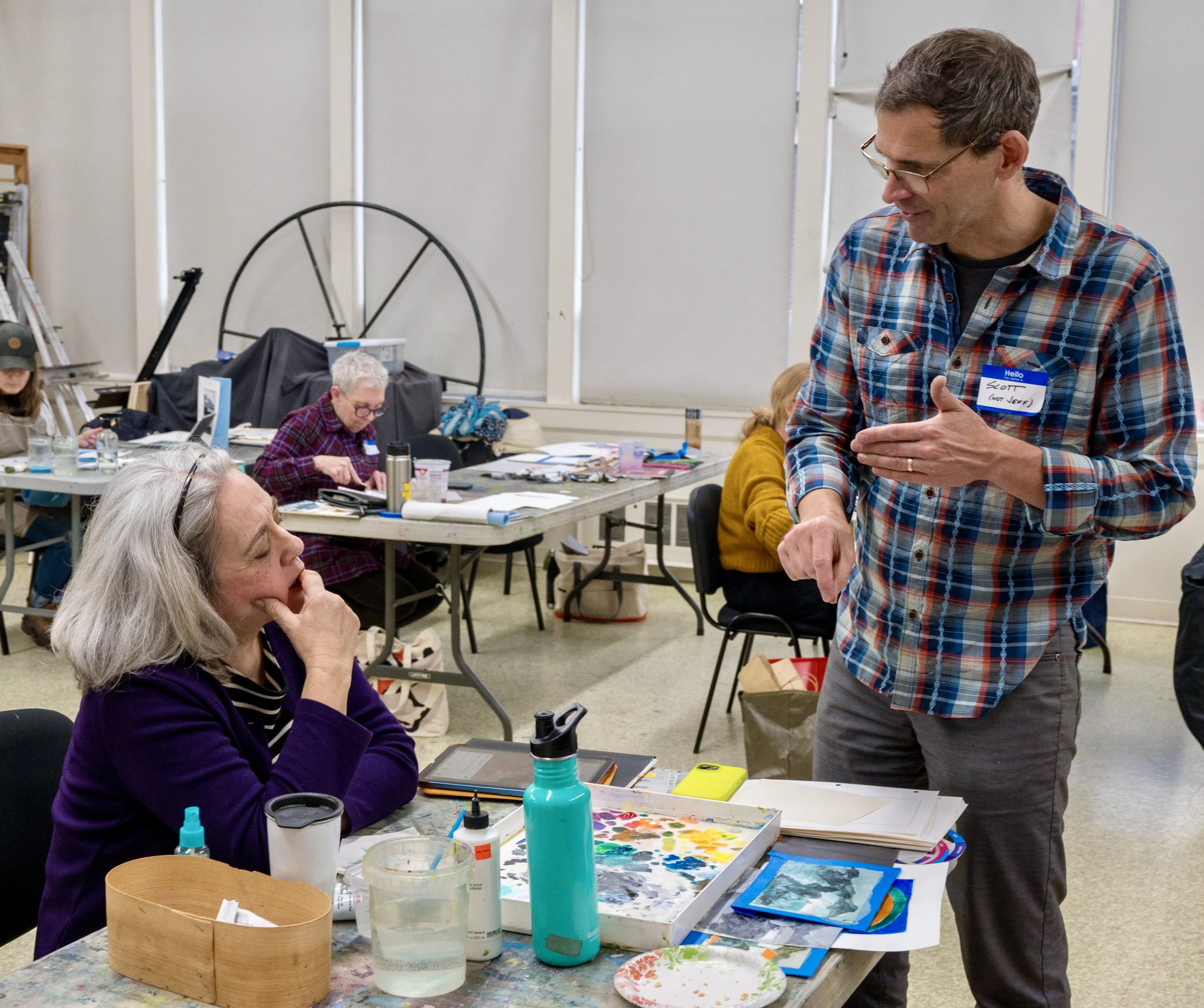 A man wearing glasses and a plaid shirt is talking to a seated woman with gray hair and a purple sweater in an art classroom. The woman is listening attentively, surrounded by art supplies on the table, including paint, brushes, and water.