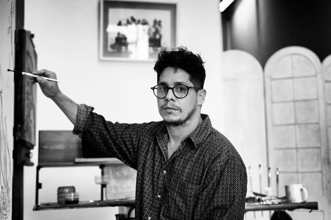 Man with glasses and dark hair in a patterned shirt painting on an easel in a room with decorative room dividers and wall art.