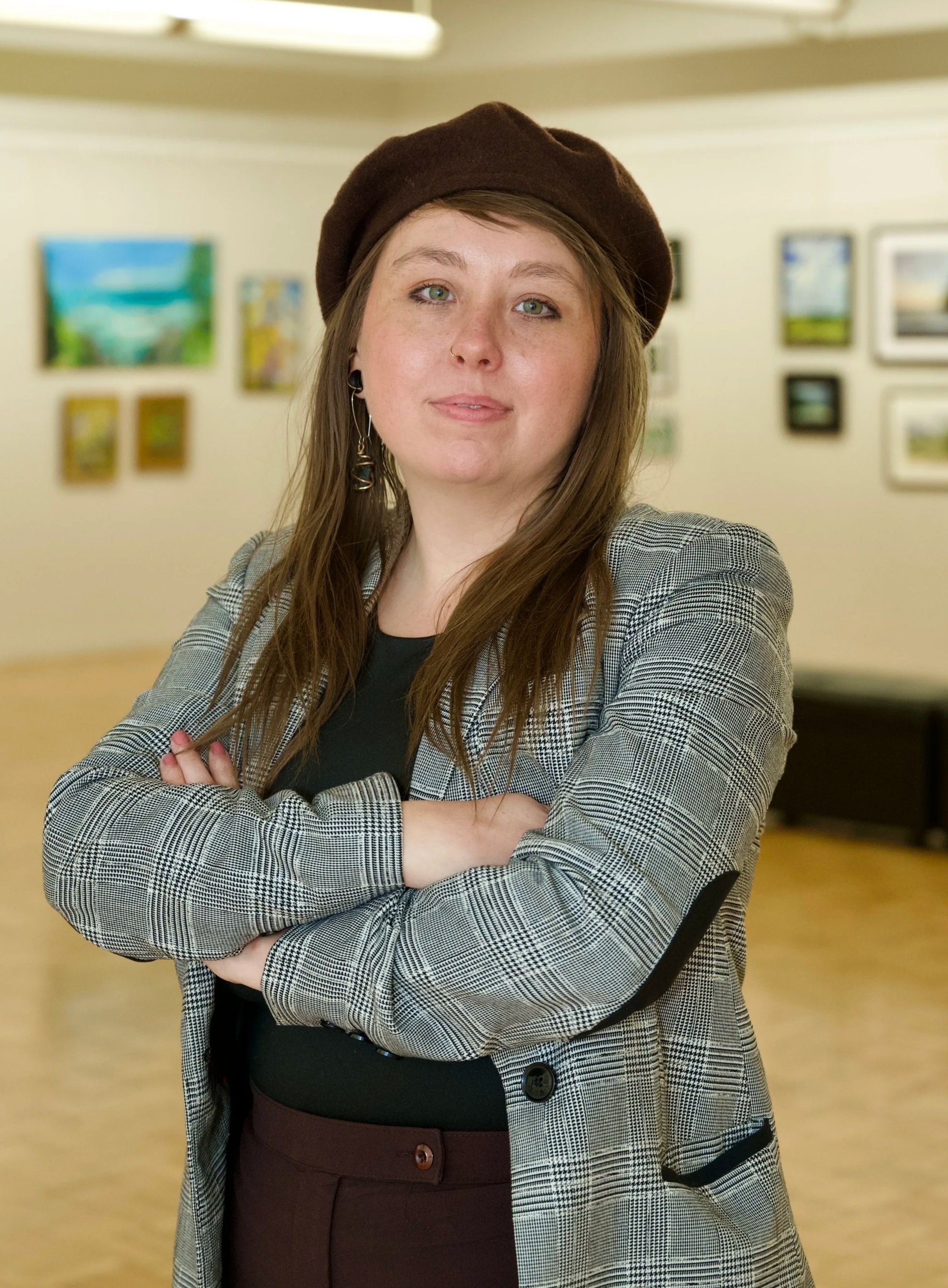A young woman with long brown hair, wearing a brown beret, a plaid blazer, and hoop earrings, standing with arms crossed in an art gallery with paintings on the wall behind her.