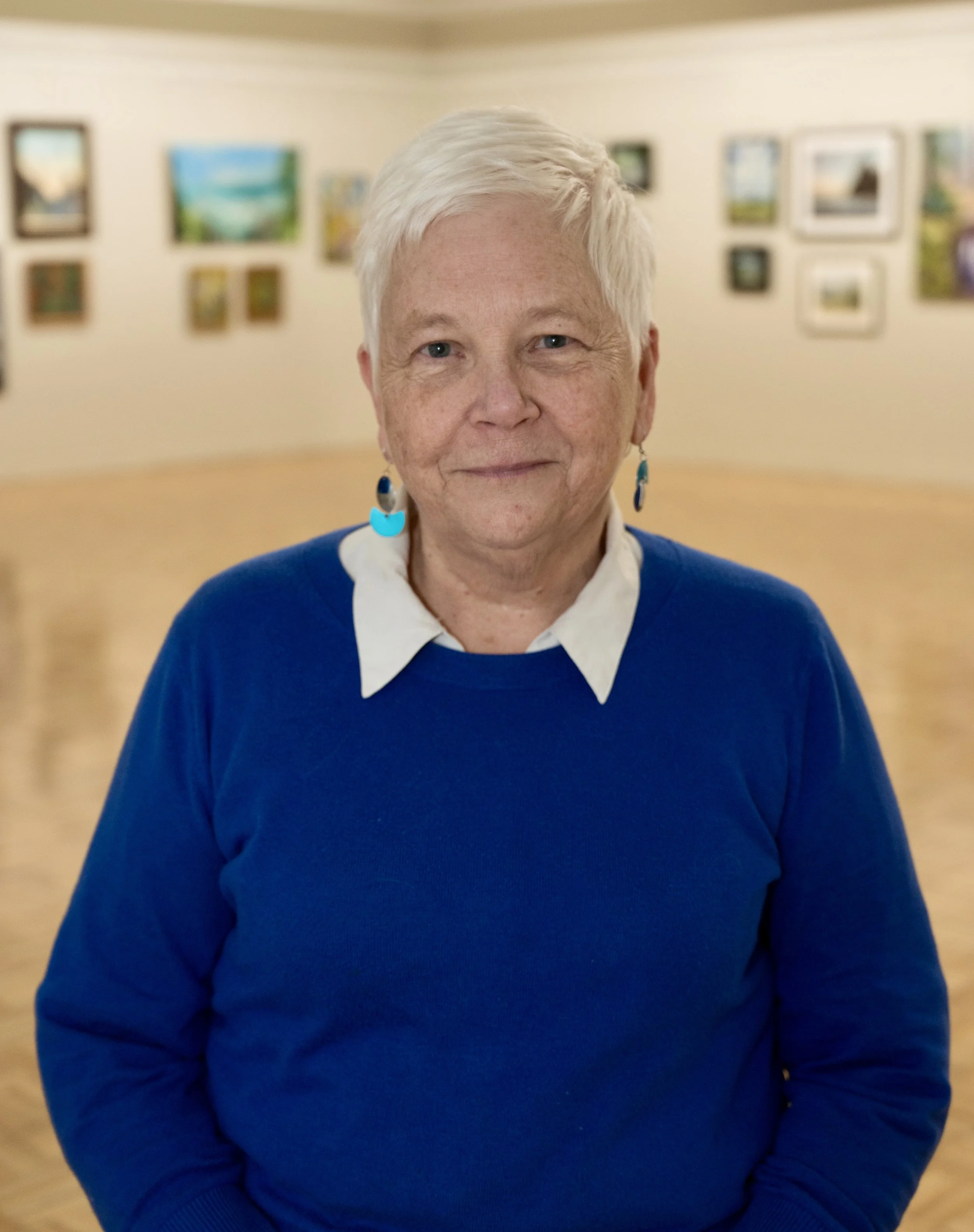Portrait of an elderly woman with short white hair, wearing a blue sweater over a white collared shirt, standing in an art gallery with various framed artworks on the walls.