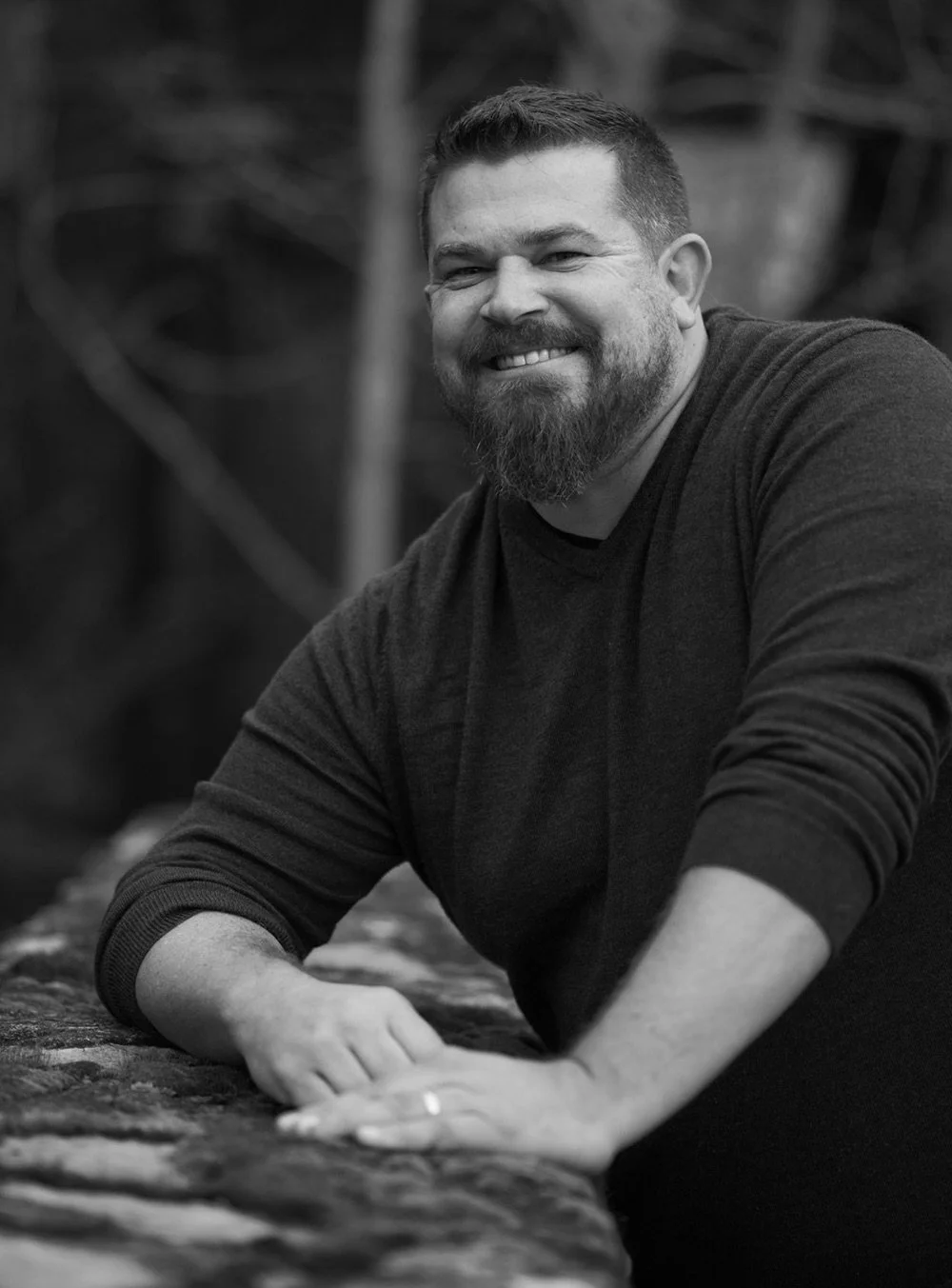A smiling man with a beard and short hair, wearing a long-sleeve shirt, leaning on a stone surface outdoors.
