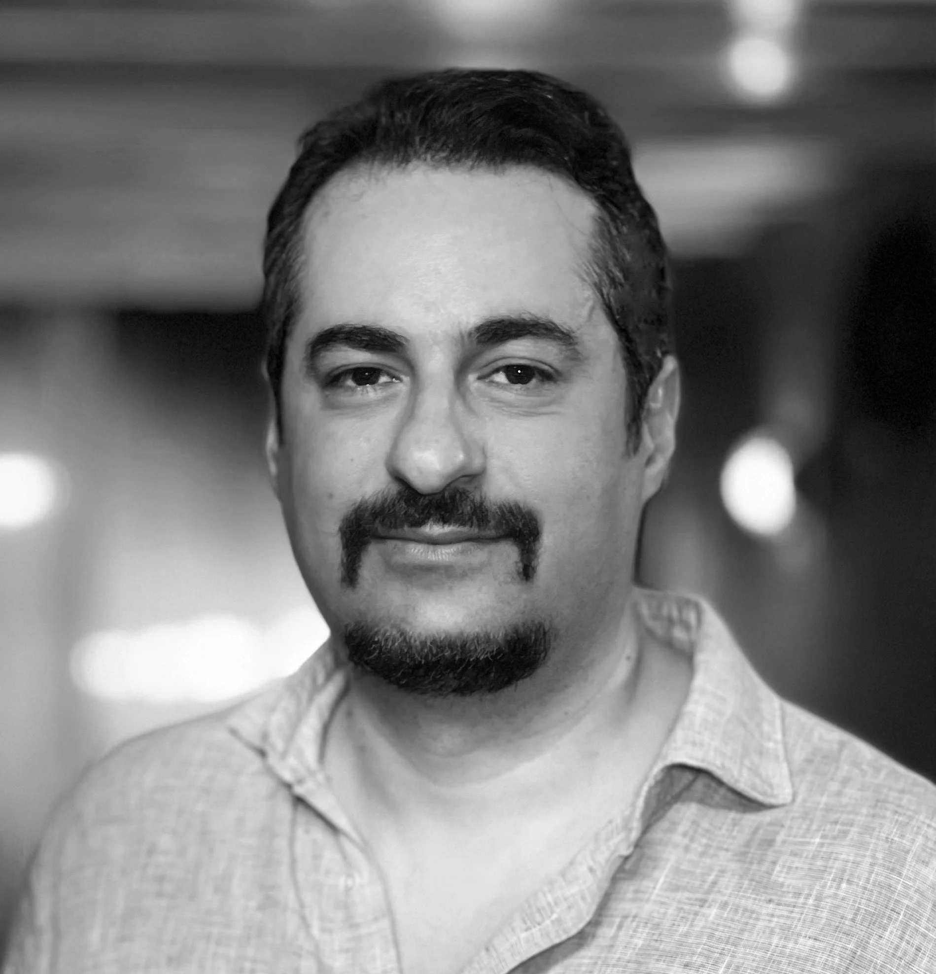 Black and white portrait of a man with dark hair, a mustache and goatee, wearing a casual button-down shirt, in an indoor setting with blurred background lights.