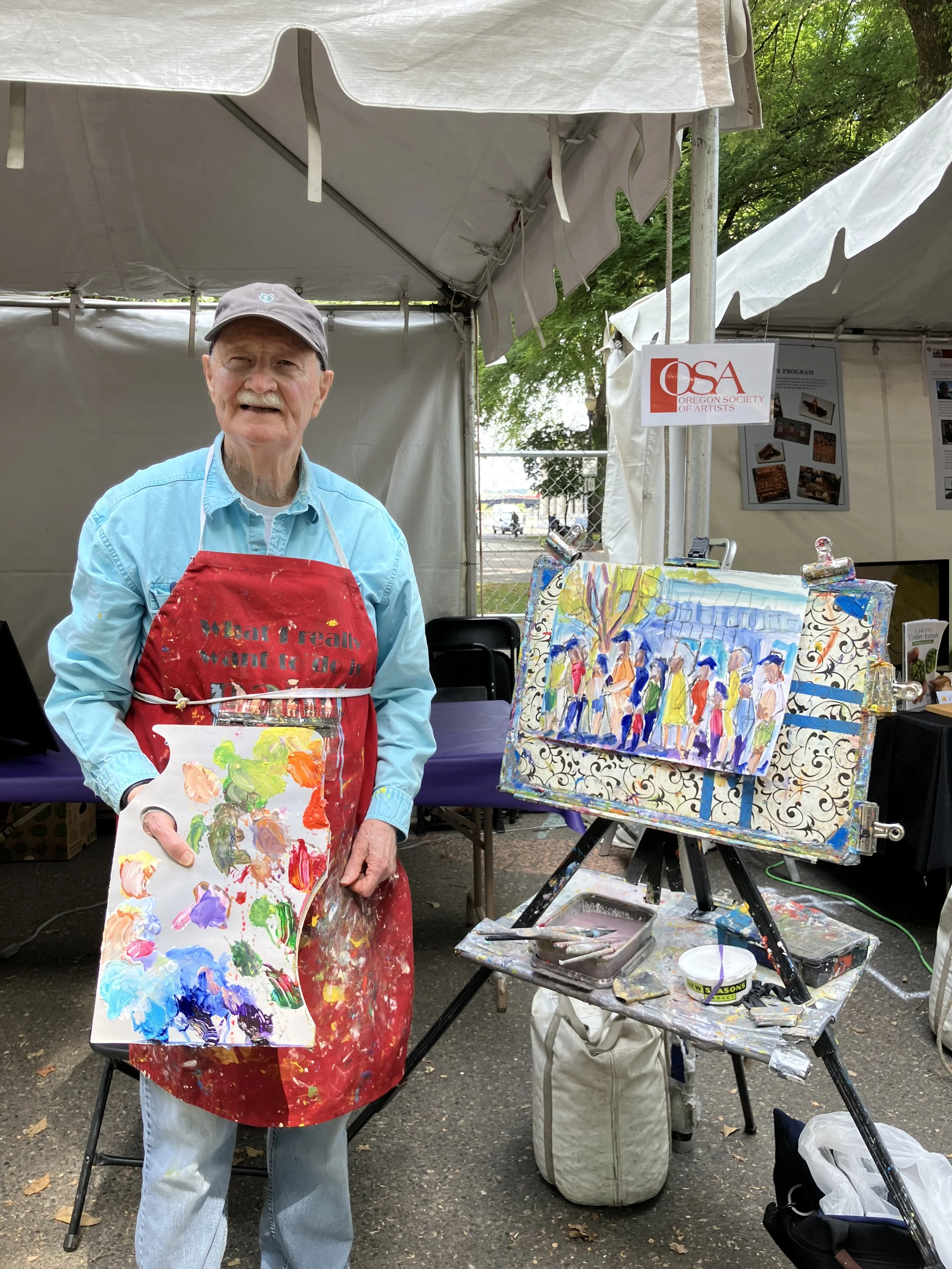 An elderly man standing in front of an artist's display at an outdoor art fair, holding a palette with colorful paints and wearing a red apron, blue shirt, and cap. There is a painting of people lined up on an easel nearby and a sign for the Oregon Society of Artists.