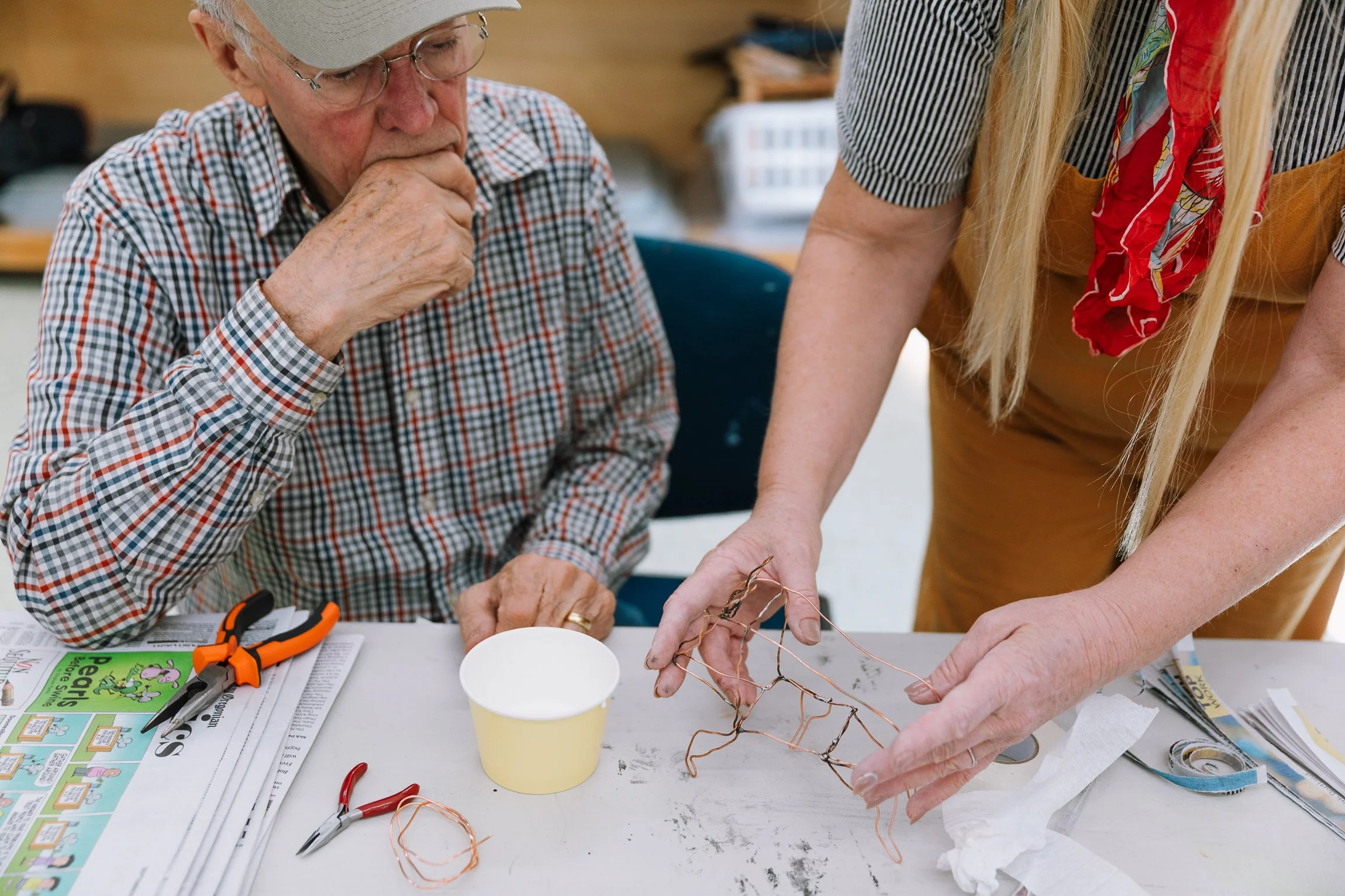 An elderly man and a woman are working on a craft project involving twisted copper wire to create a wire sculpture. The man is observing carefully, resting his chin on his hand, while the woman is holding the wire structure. There are scissors, a yellow cup, and a newspaper on the table.