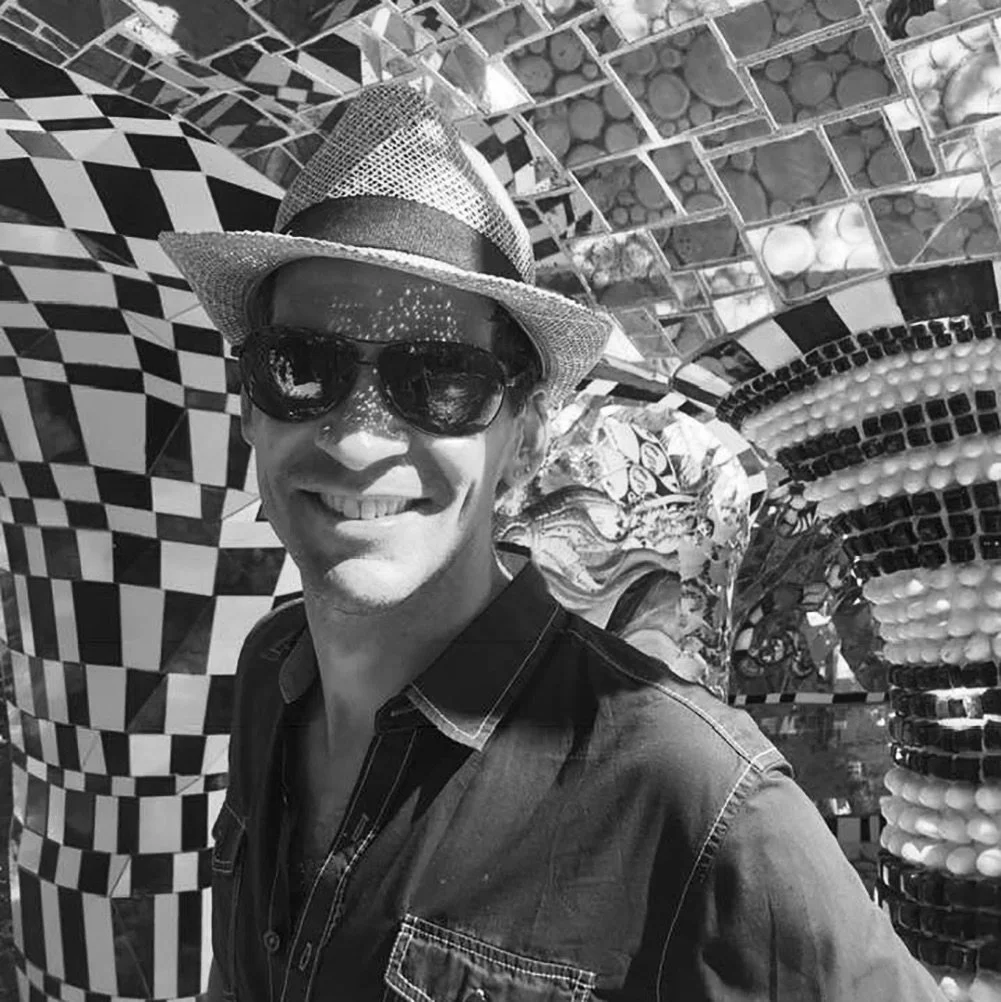 A smiling man wearing sunglasses, a hat, and a denim shirt, standing in front of an optical illusion artwork with checkered and spiral patterns.