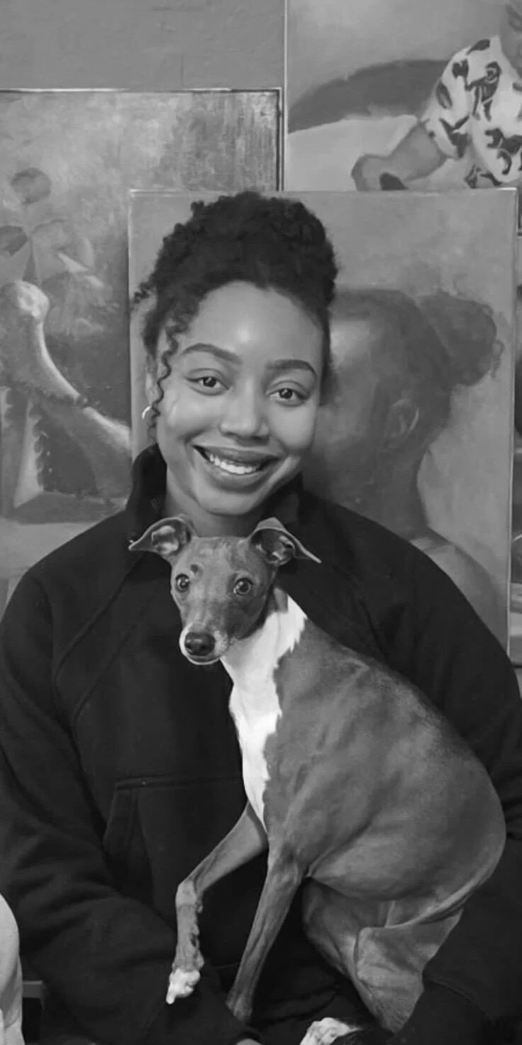 A smiling woman with her hair in a puff hairstyle, wearing a dark jacket, is holding a Greyhound dog with a white chest and a short coat. The background features artwork with visible paintings.