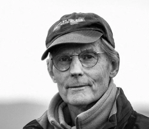 A smiling man with glasses and spiky hair in an art studio, with paintings of landscapes in the background.