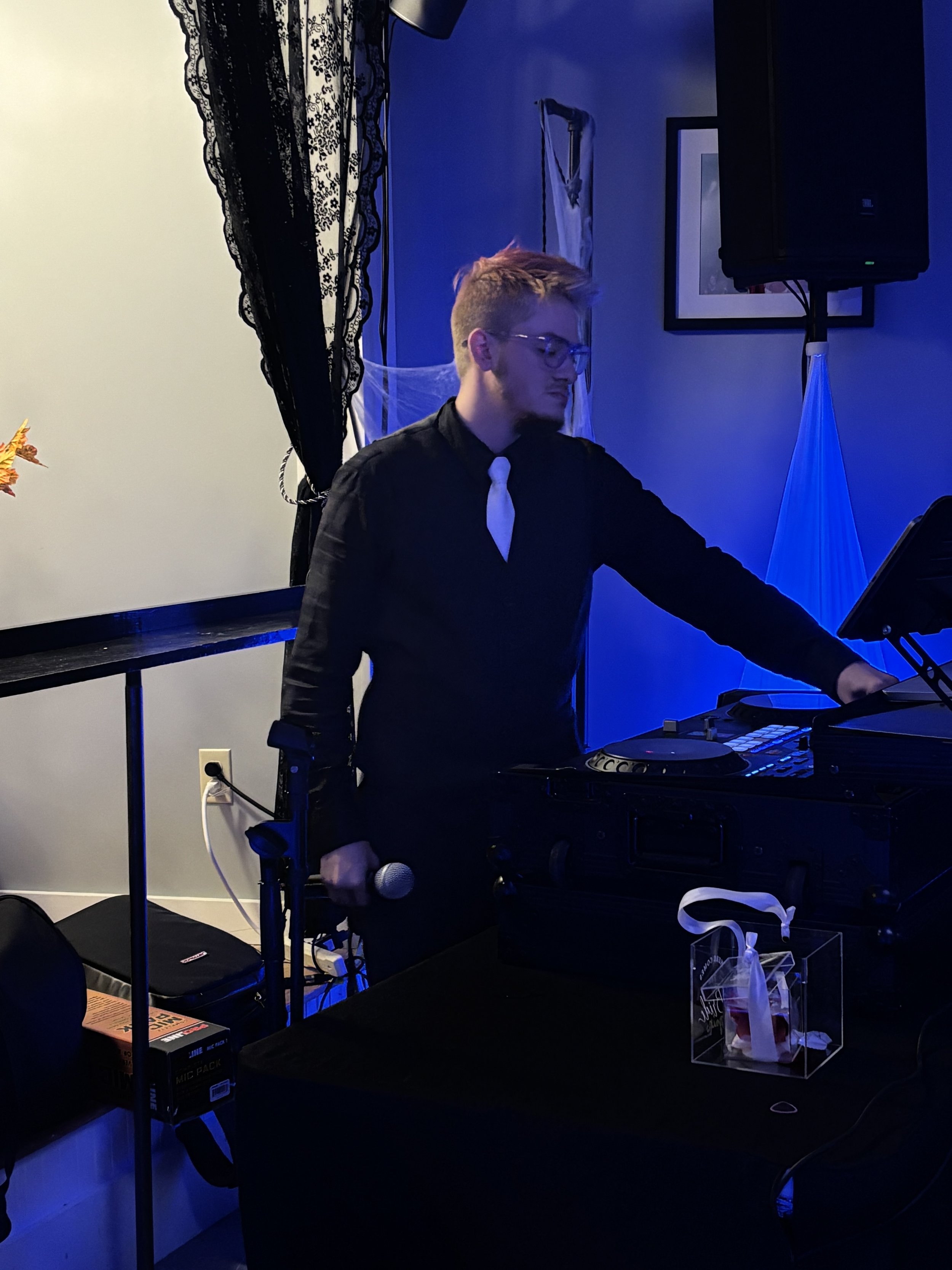 A young man with glasses and blonde hair dressed in a black shirt and white tie, DJing at a party or event with DJ equipment, microphone, and blue lighting.