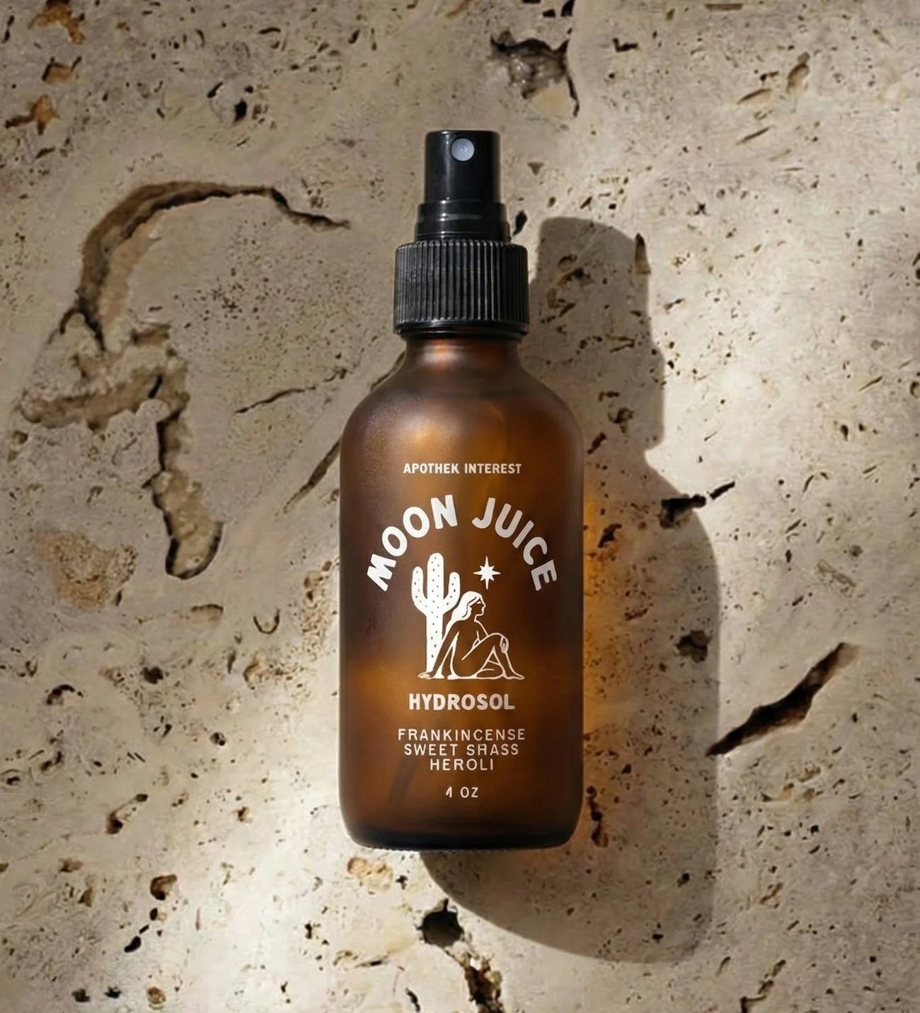 Top-down view of Apothek Interest's Moon Juice botanical face mist in an amber glass bottle, lying on a natural travertine stone surface in the sunlight.