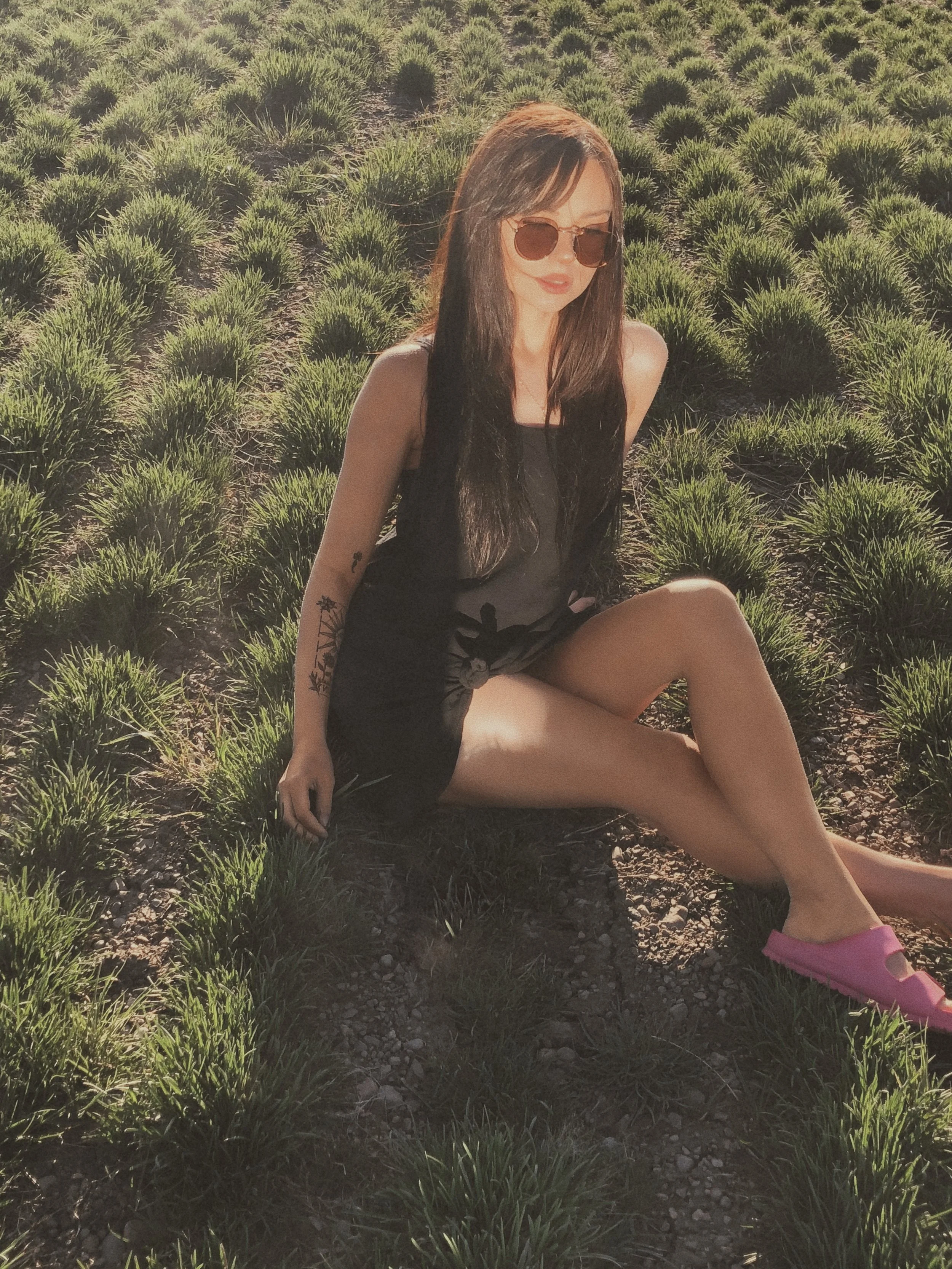The founder of Apothek Interest captures a moment of earthy repose in a sunlit landscape, wearing minimal aesthetic outfit and sunglasses.