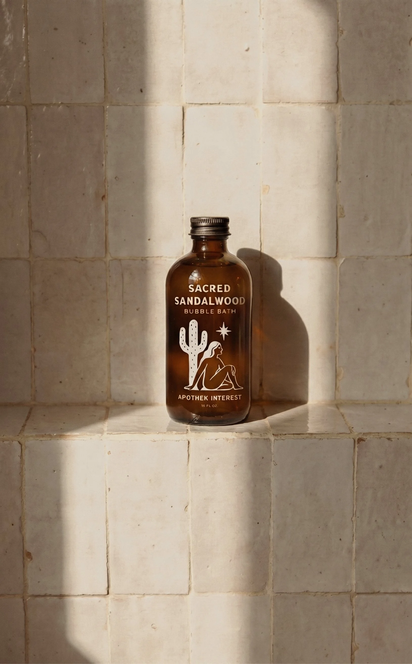 16 oz amber glass bottle of Apothek Interest Sacred Sandalwood Bubble Bath on a sunlit neutral tiled bathroom shelf