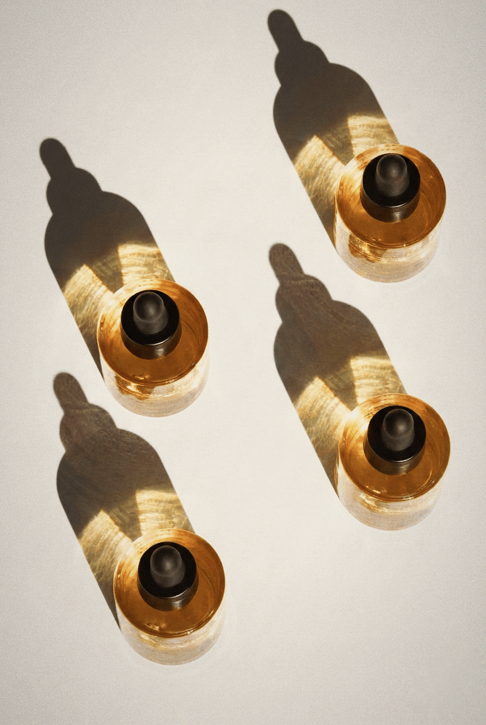 Top-down view of four Apothek Interest Solar Face Oil 1 oz dropper bottles. Sunlight shines through the golden botanical desert serum, casting long, dramatic shadows on a light surface.