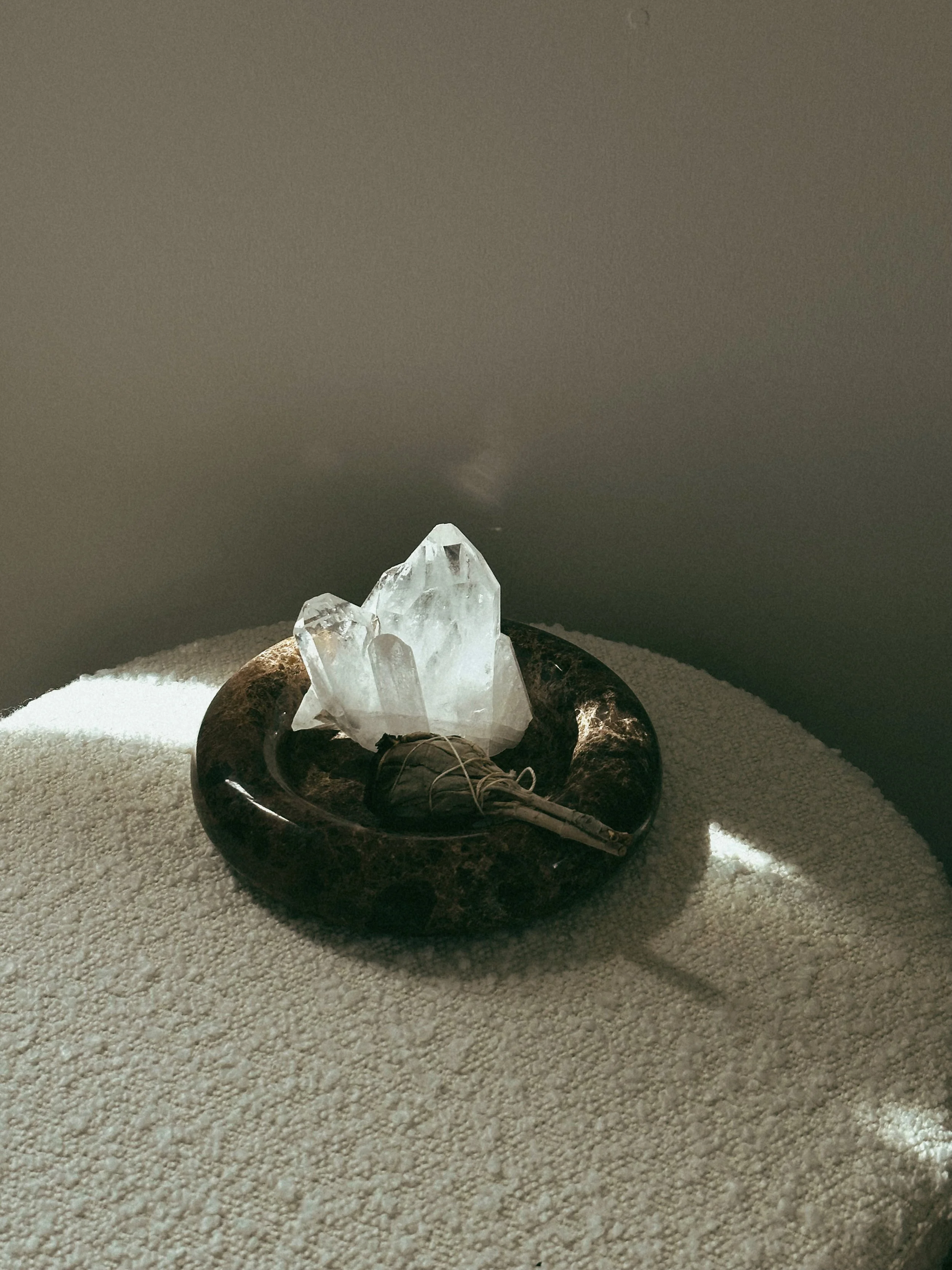 A raw clear quartz crystal cluster and a tied smudge bundle resting in a dark stone bowl in natural sunlight, representing an Apothek Interest ritual kit.