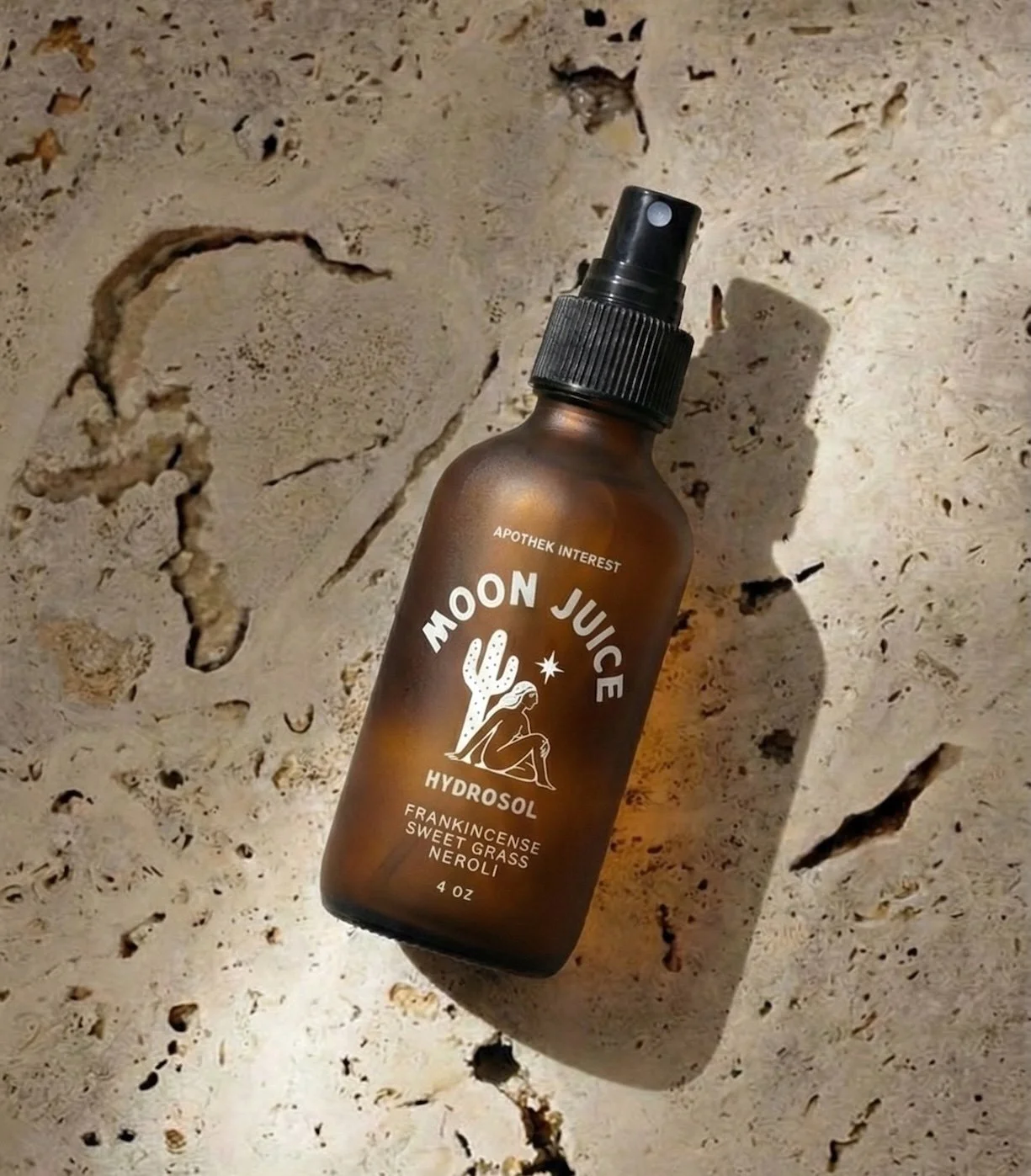 4oz amber glass bottle of Moon Juice Hydrosol face mist by Apothek Interest, resting diagonally on a sunlit, textured earthy stone surface.