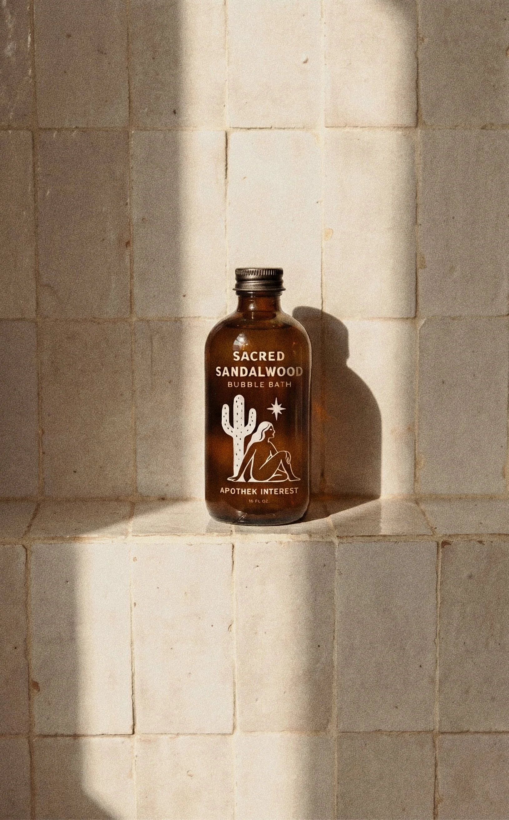 Amber glass bottle of Apothek Interest Sacred Sandalwood Bubble Bath resting on warm, sunlit cream tiles.