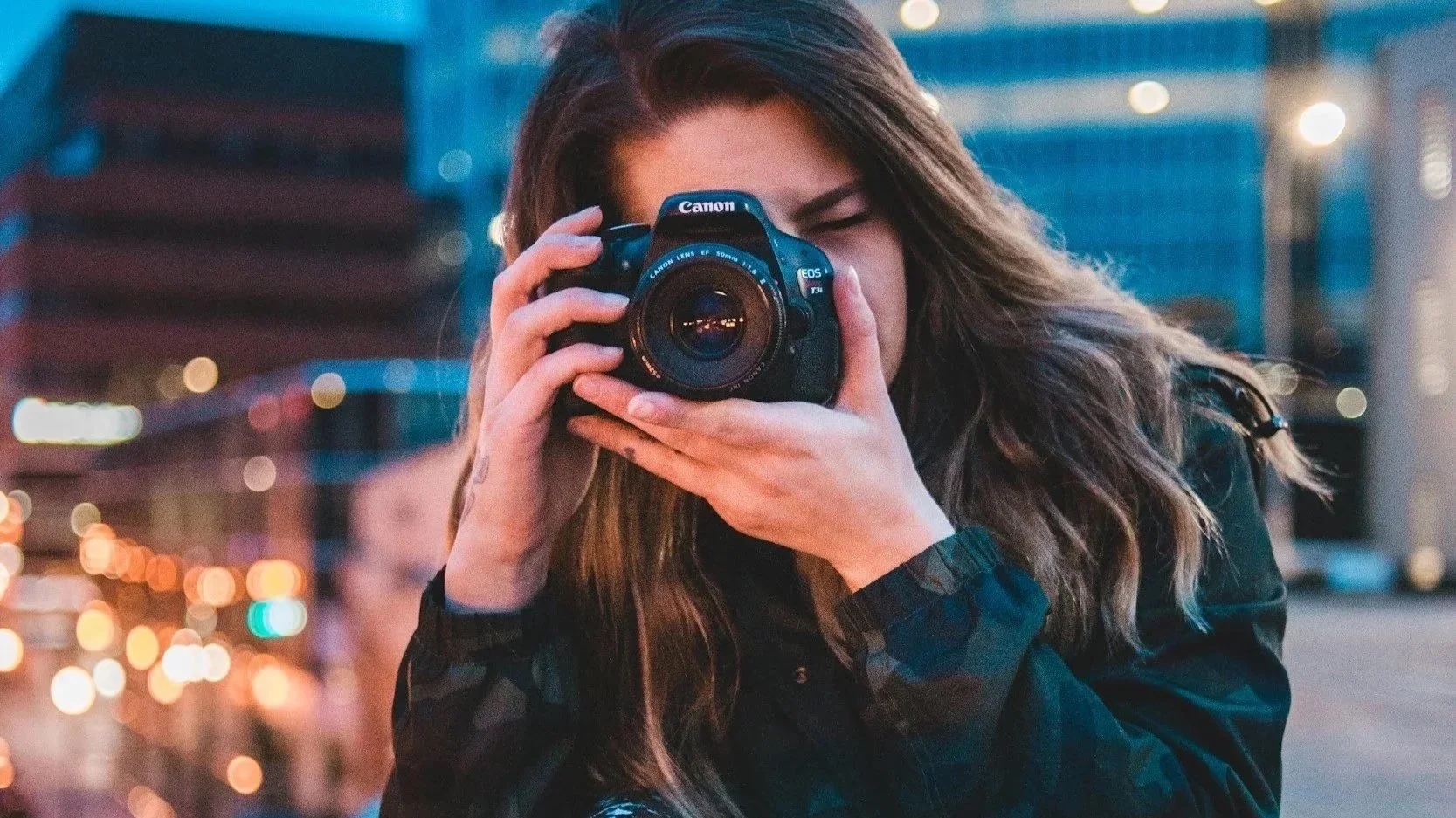 A woman with long hair holding a Canon DSLR camera up to her face, appearing to take a photo, in an urban setting during dusk with blurred city lights in the background.