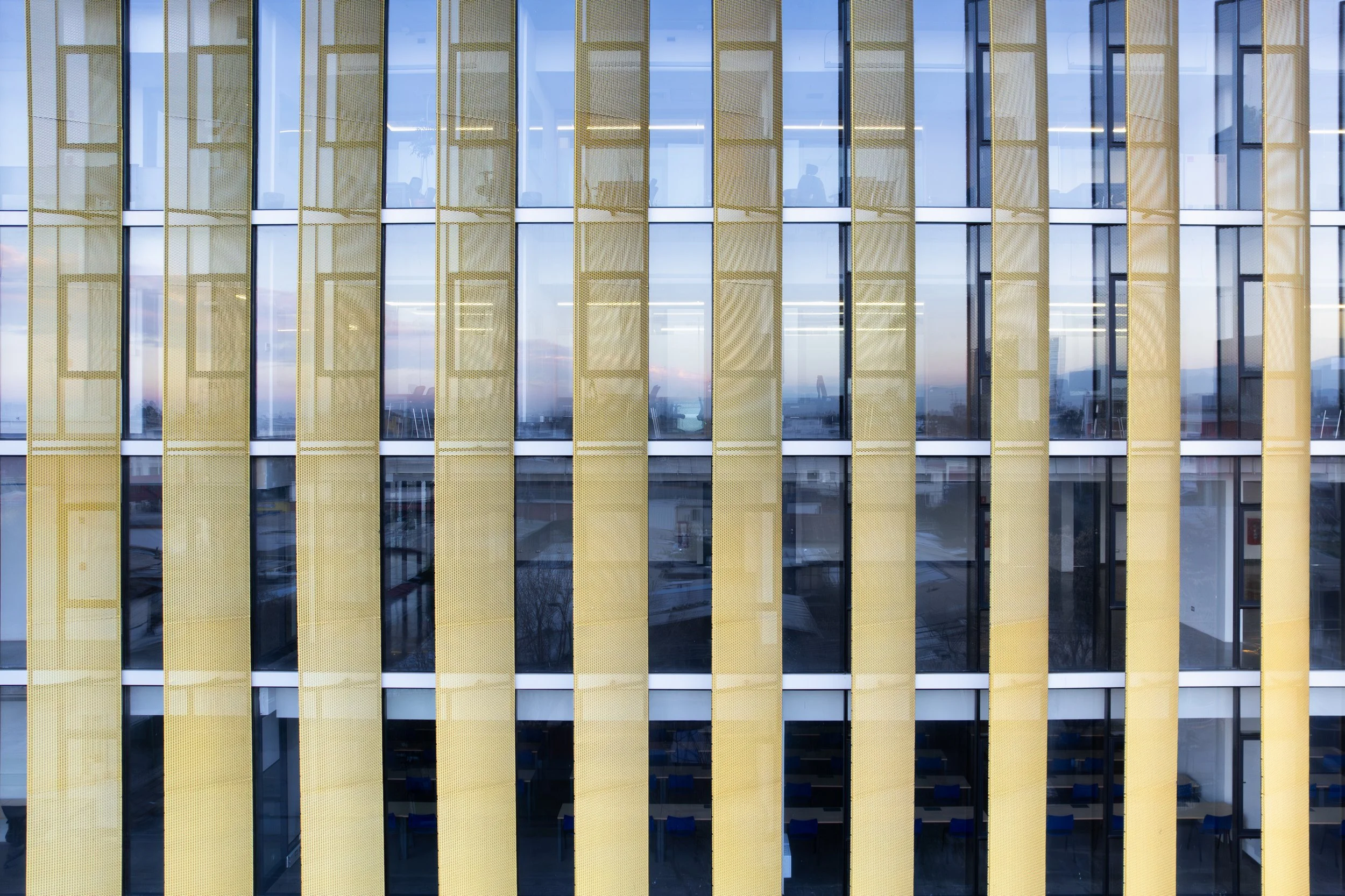 Fachada de un edificio moderno con ventanas de vidrio y paneles en metal desplegado anodizado color oro.