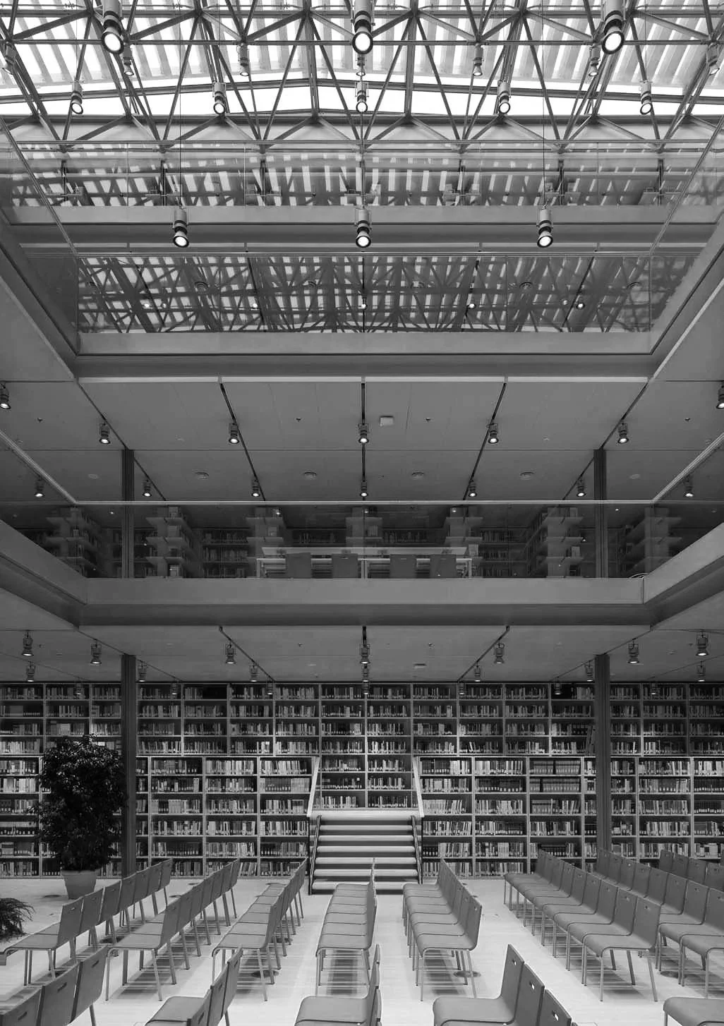 Interior de una biblioteca moderna con estanterías llenas de libros, área de lectura con sillas alineadas y un acceso por escaleras en el Fondo.