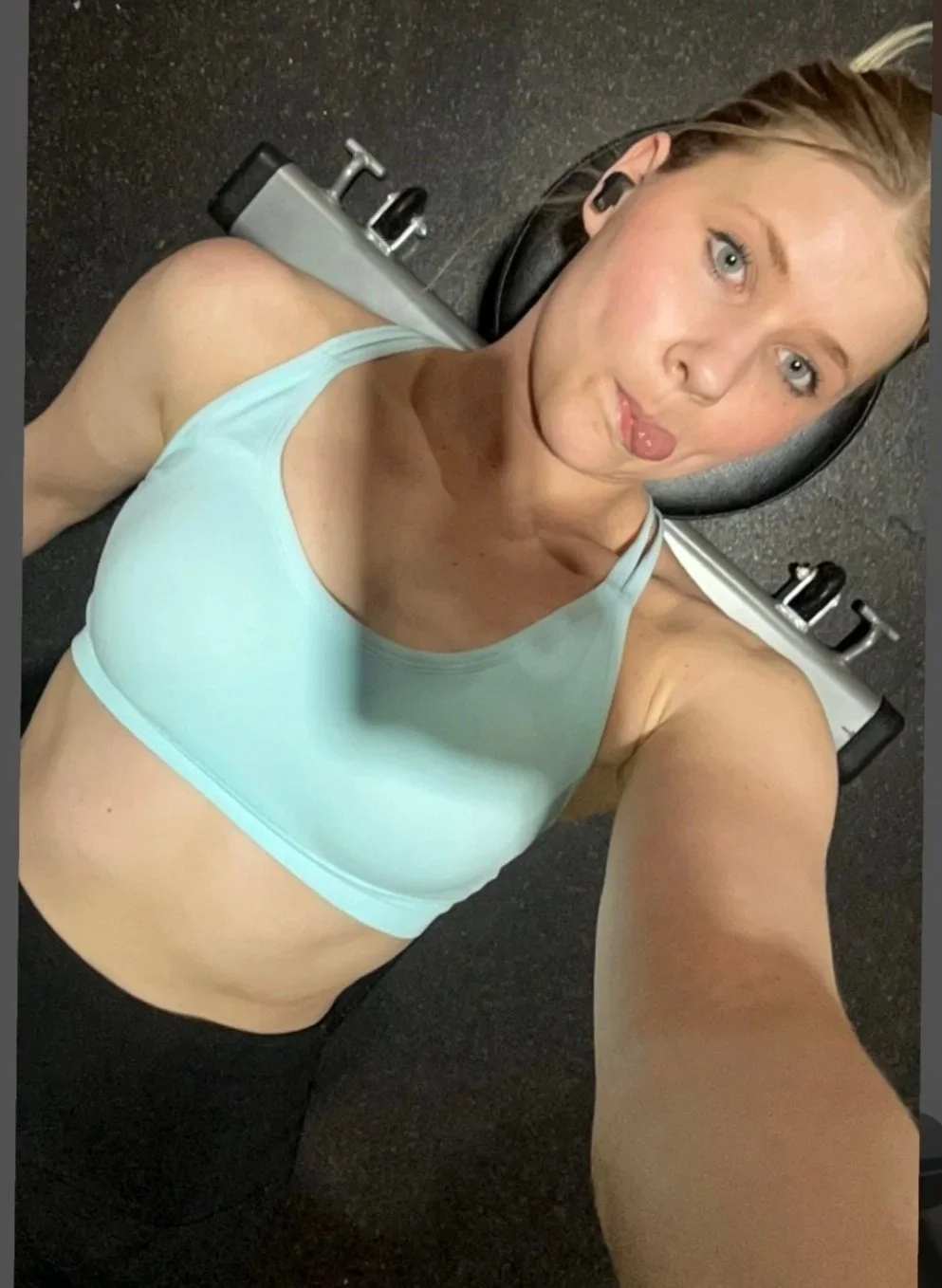 A woman lying on her back on a gym floor, taking a selfie. She is wearing a light blue sports bra and black workout pants. There is gym equipment behind her, including a weight bench.