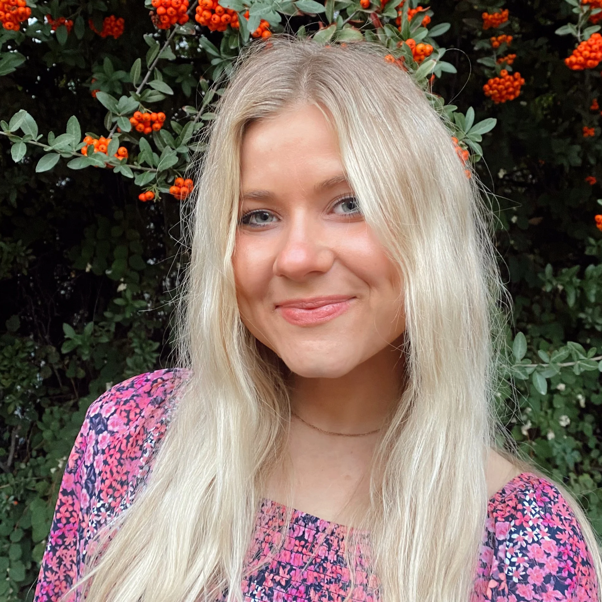 A young woman with long blonde hair and blue eyes, smiling, standing in front of green foliage with orange berries, wearing a pink and purple patterned top.