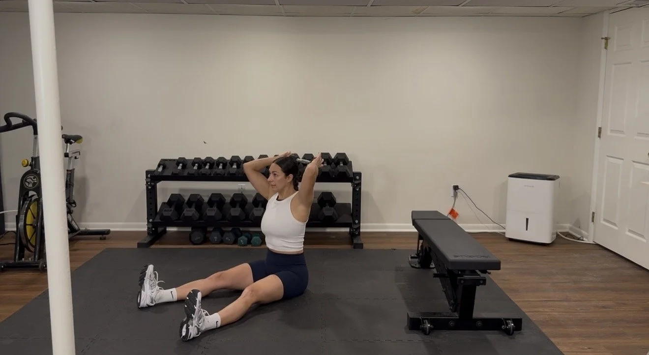 A woman performing sit-up exercises on a workout mat in a home gym with dumbbells, a bench, and exercise equipment in the background.