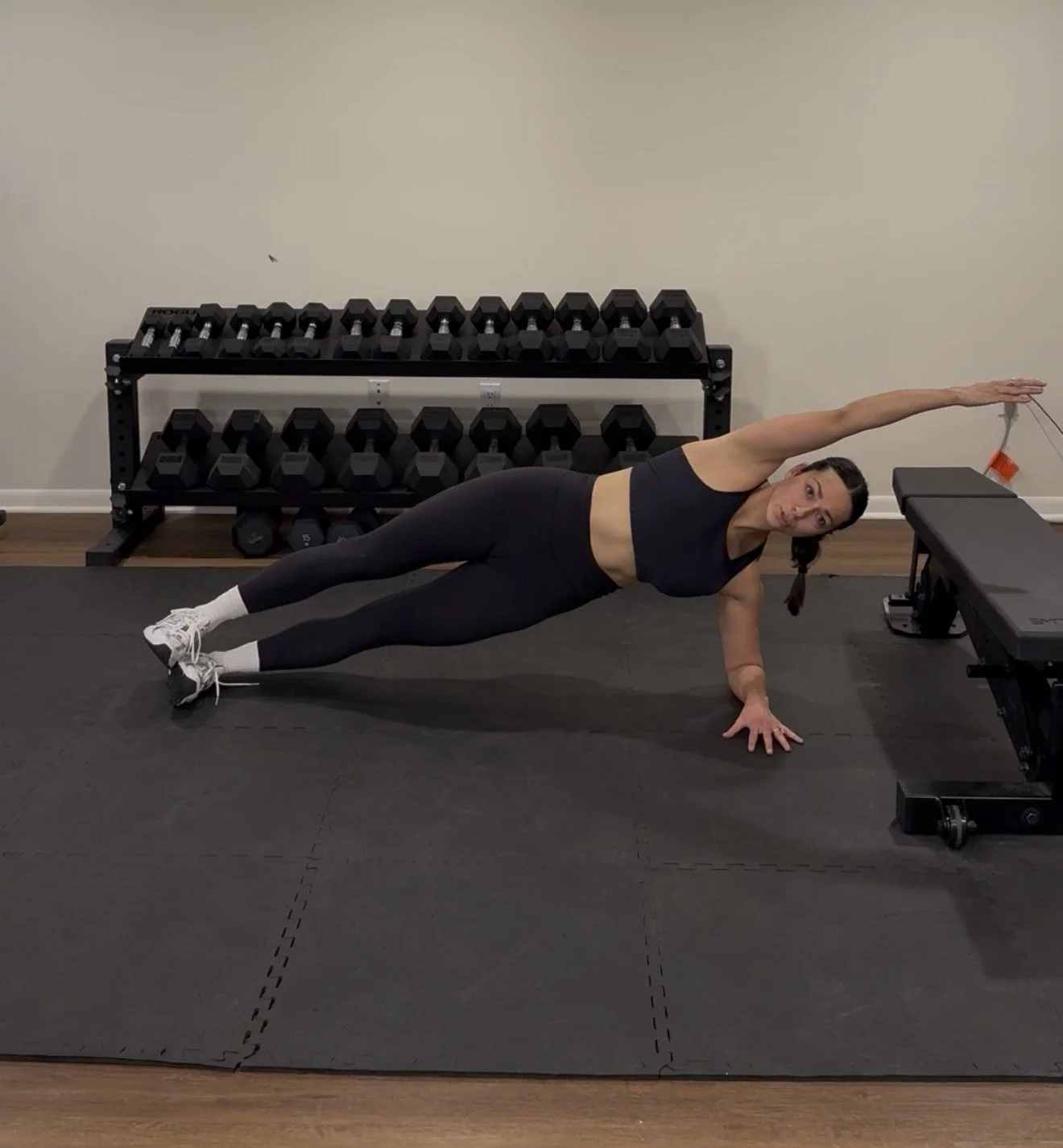 A woman performing a side plank exercise on a black exercise mat in a gym, reaching her right arm over her head, with a rack of dumbbells in the background.