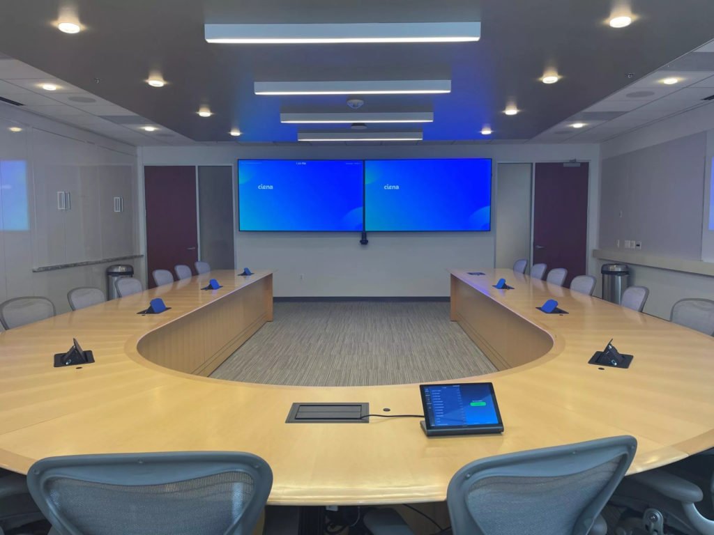A modern conference room with a U-shaped wooden table set with microphones, blue notepads, and a tablet. The room has gray chairs, a large video display screen on the wall, and overhead lighting.