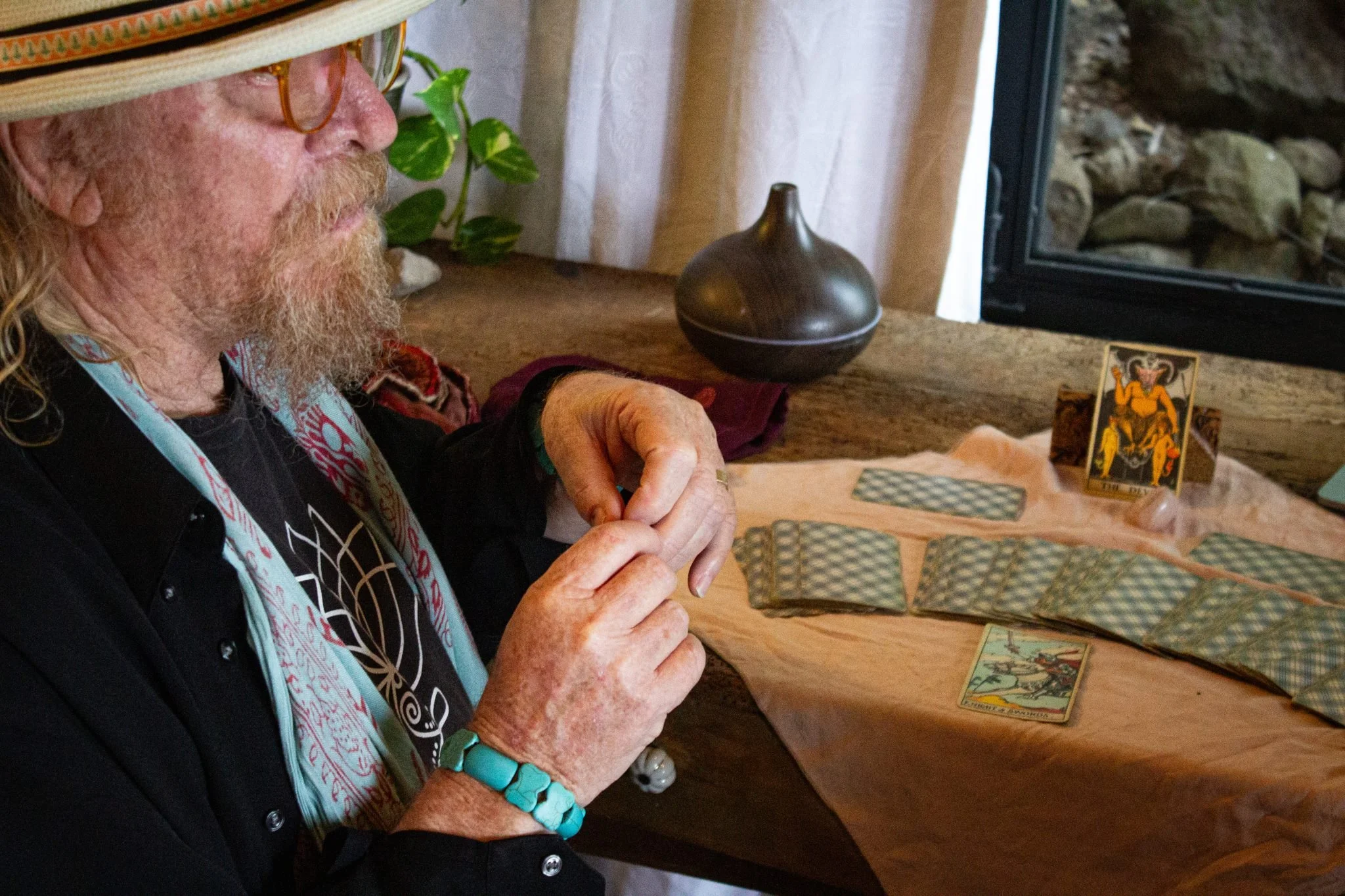 A man with long hair, glasses, and a beard wearing a patterned scarf and black shirt is sitting at a table with tarot cards and crystals, near a window.