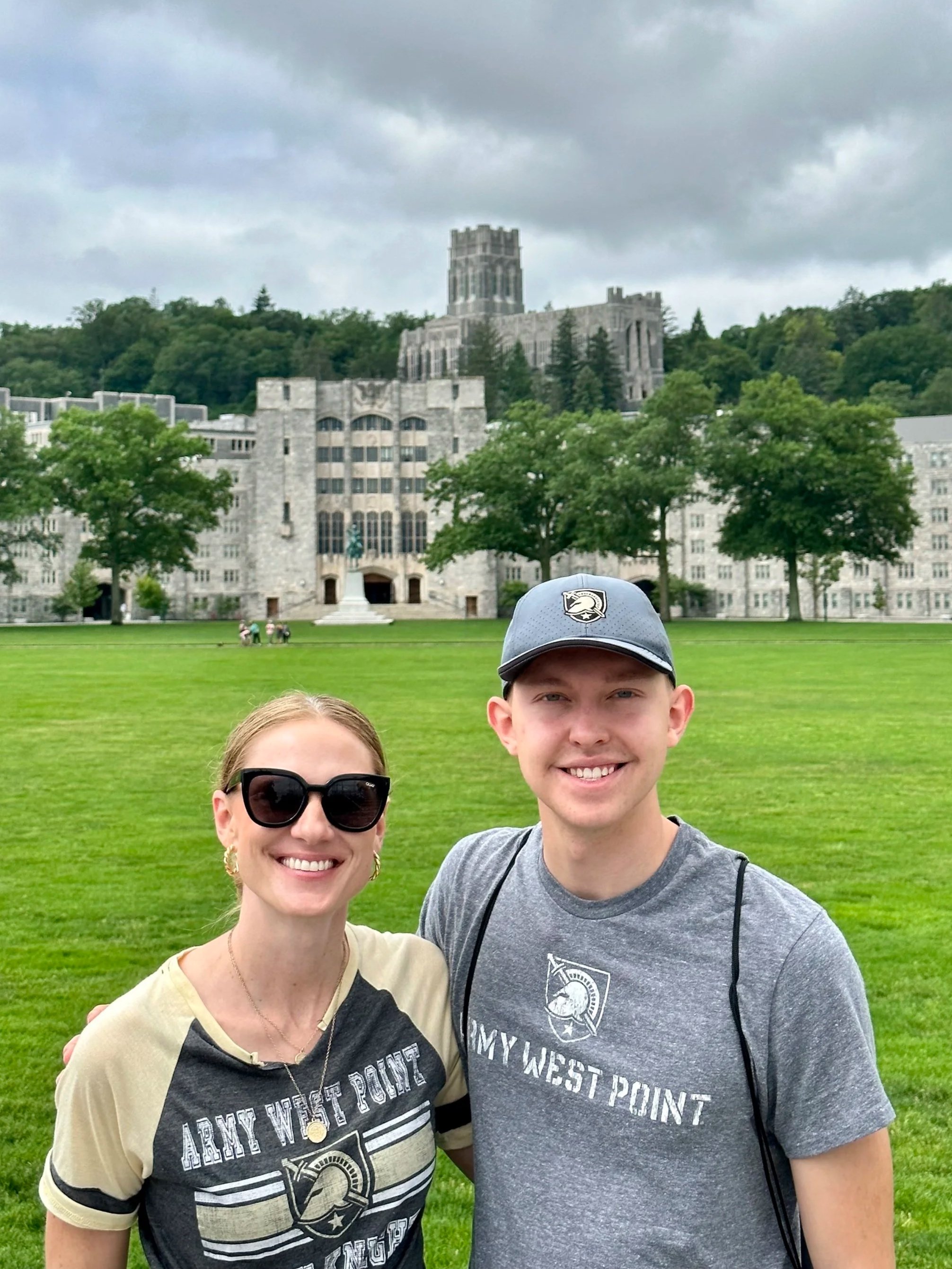 Two people smiling for a photo on a grassy field with a large stone building and trees in the background, under cloudy skies.