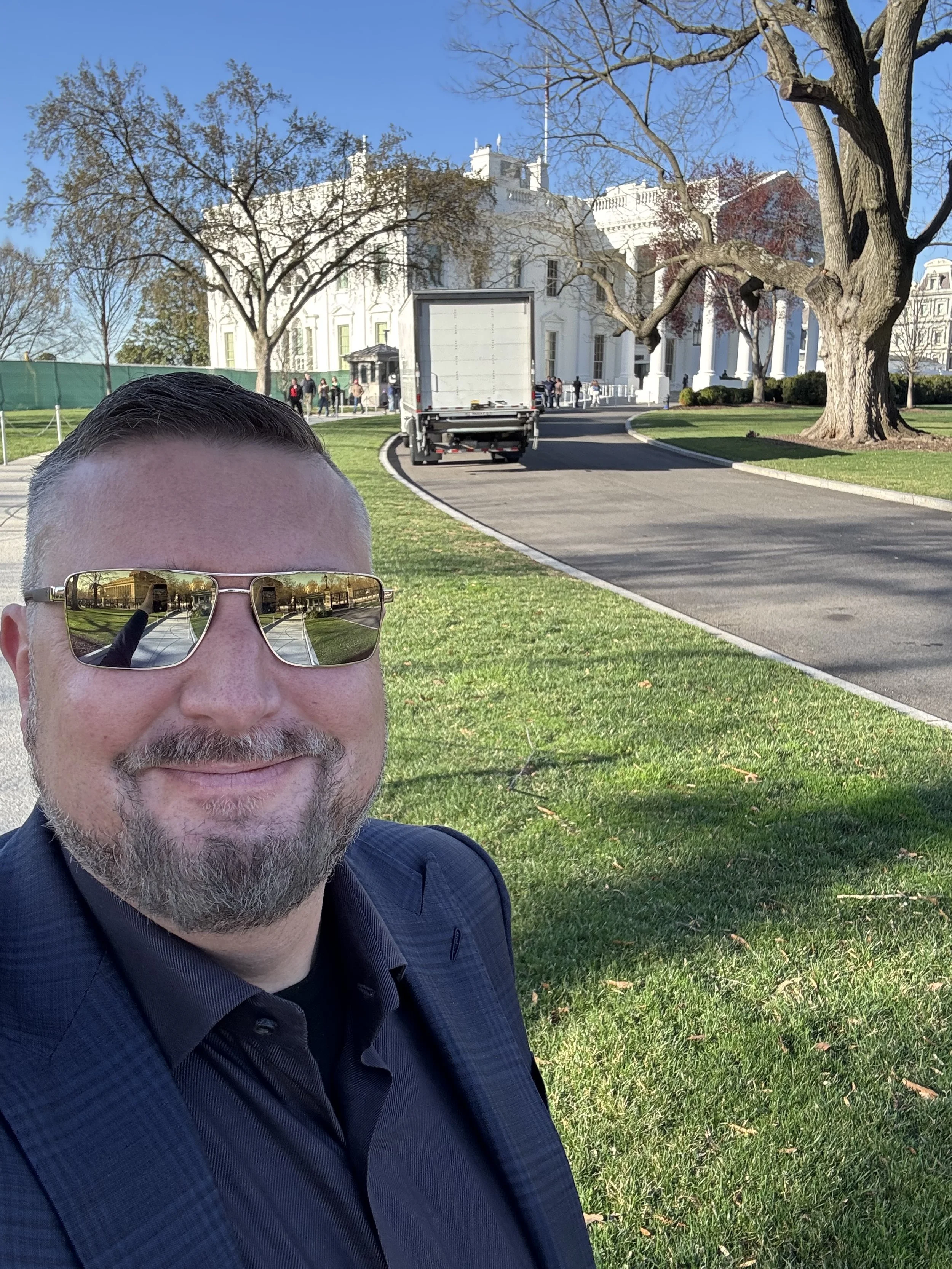 Smiling man with sunglasses taking a selfie in front of the White House, with trees, a lawn, and a truck in the background.