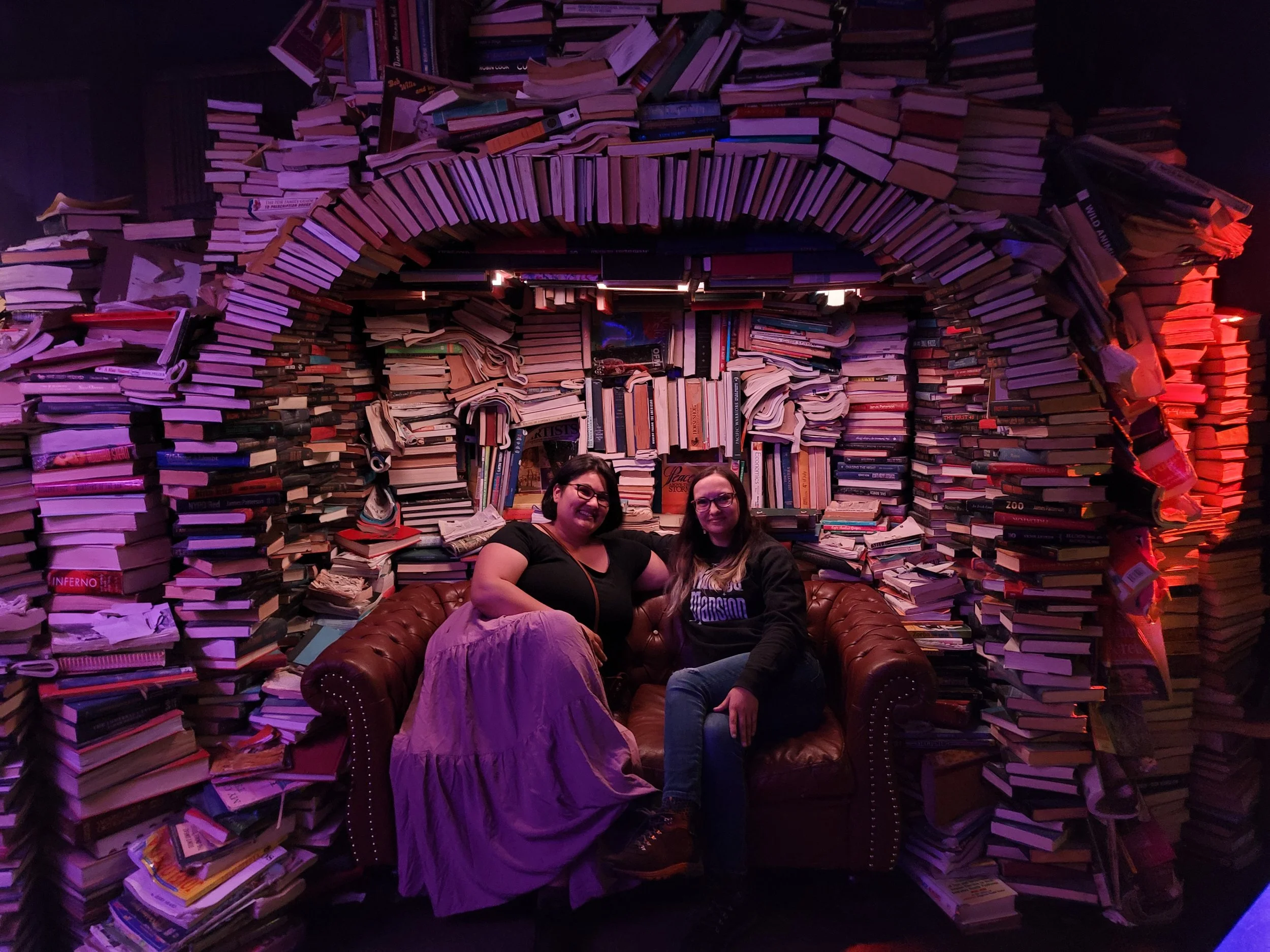 Two women sitting on a brown leather sofa in front of a wall made of books. Books are stacked and arranged to create an archway and a surrounding wall. The scene is illuminated with purple and red lighting.