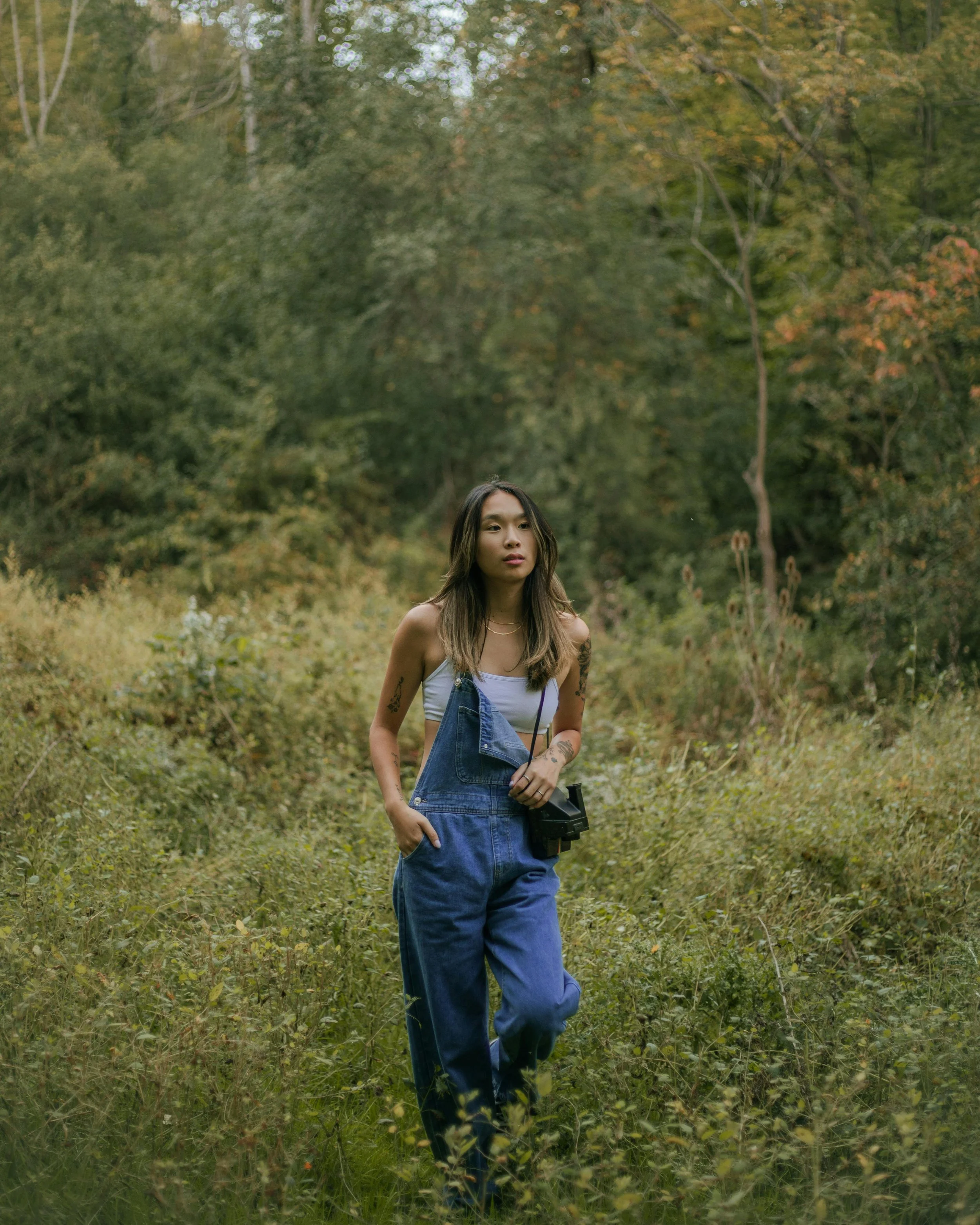 A young woman walks through a lush, green forest with autumn leaves, wearing denim overalls, a white crop top, and holding a camera in her right hand.