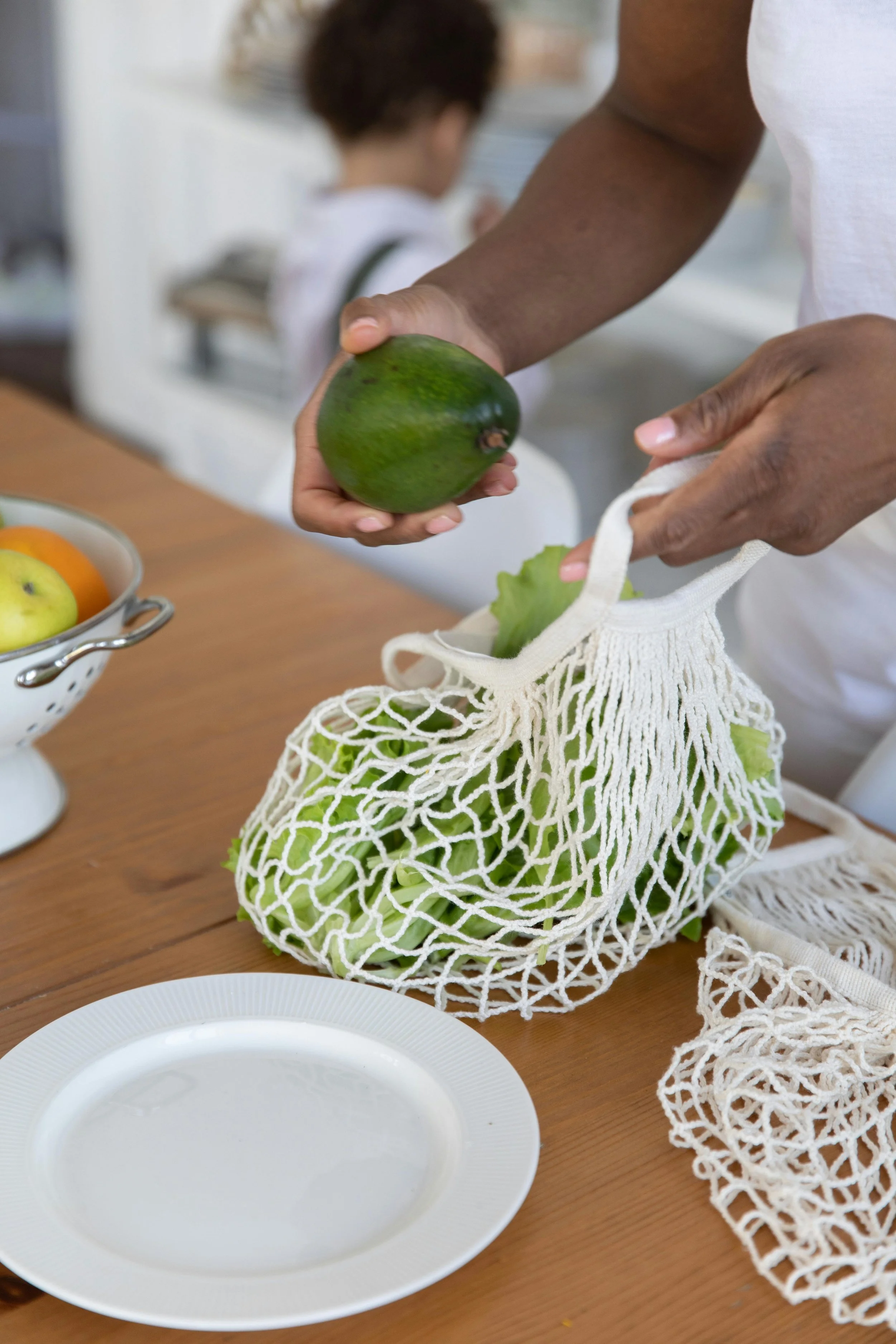 A person is holding an avocado over a reusable mesh bag filled with lettuce leaves, on a wooden table with a white plate and a bowl of apples in the background.