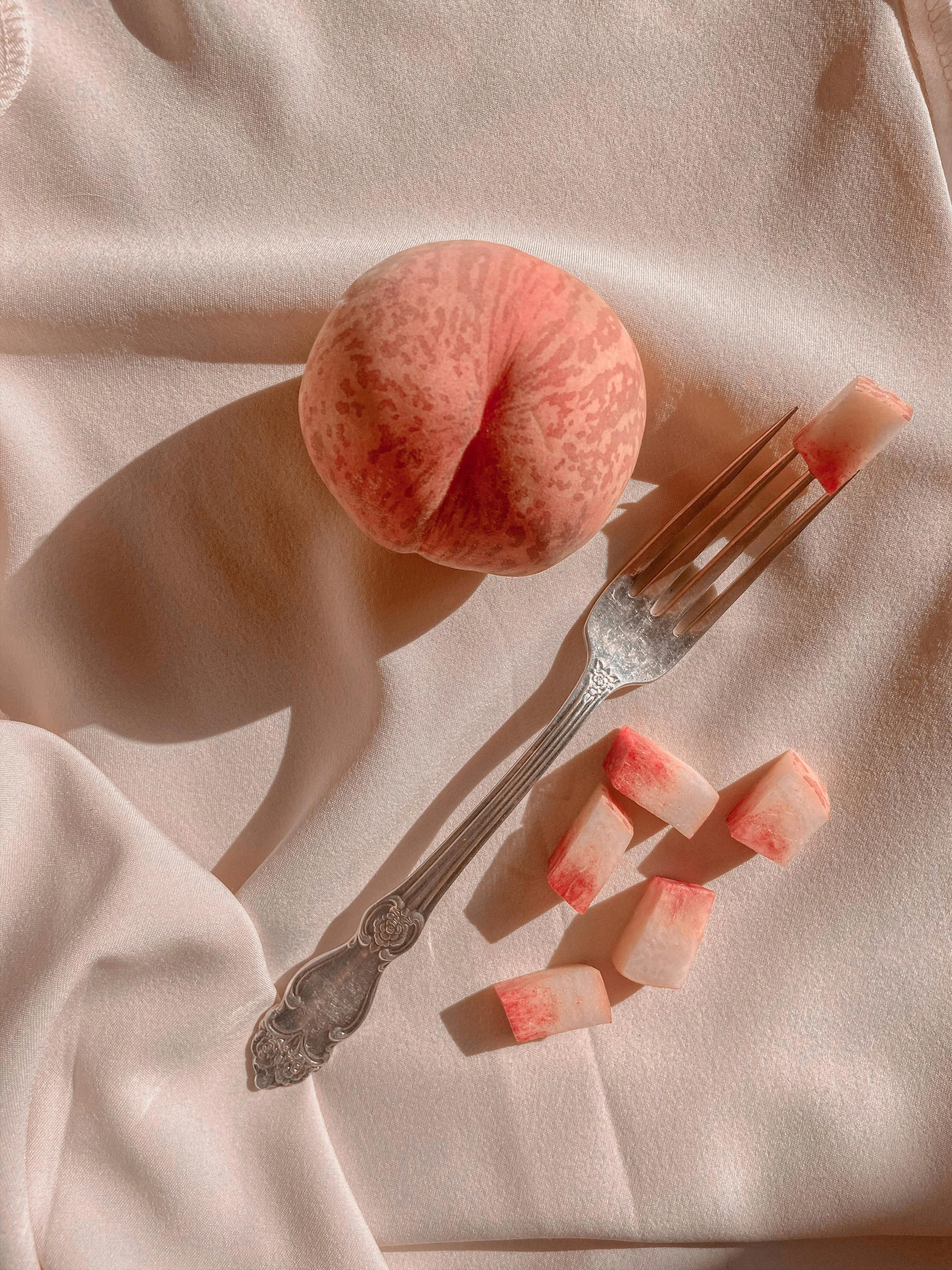 Close-up of a peeled peach, a fork with a piece of peach on it, and several small chunks of peach on a soft pink satin fabric.