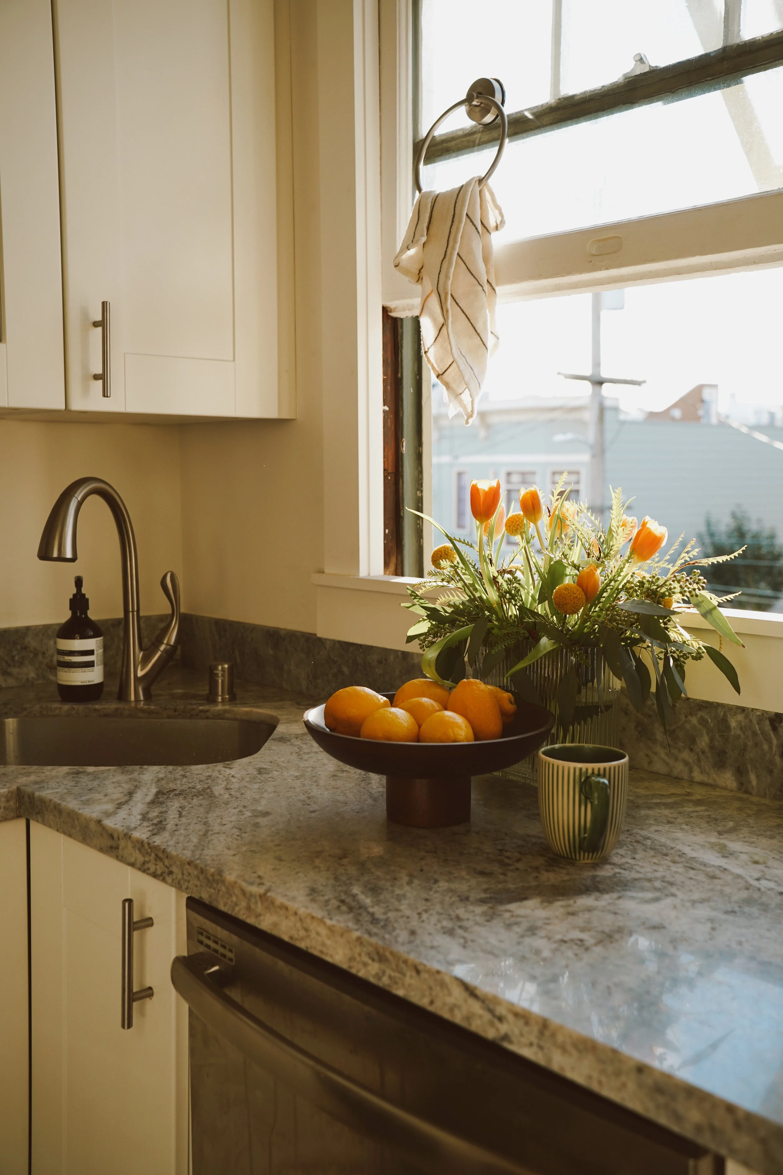 A kitchen countertop with a bowl of oranges, a bouquet of flowers, a striped mug, and a soap dispenser, with a window and cabinets in the background.