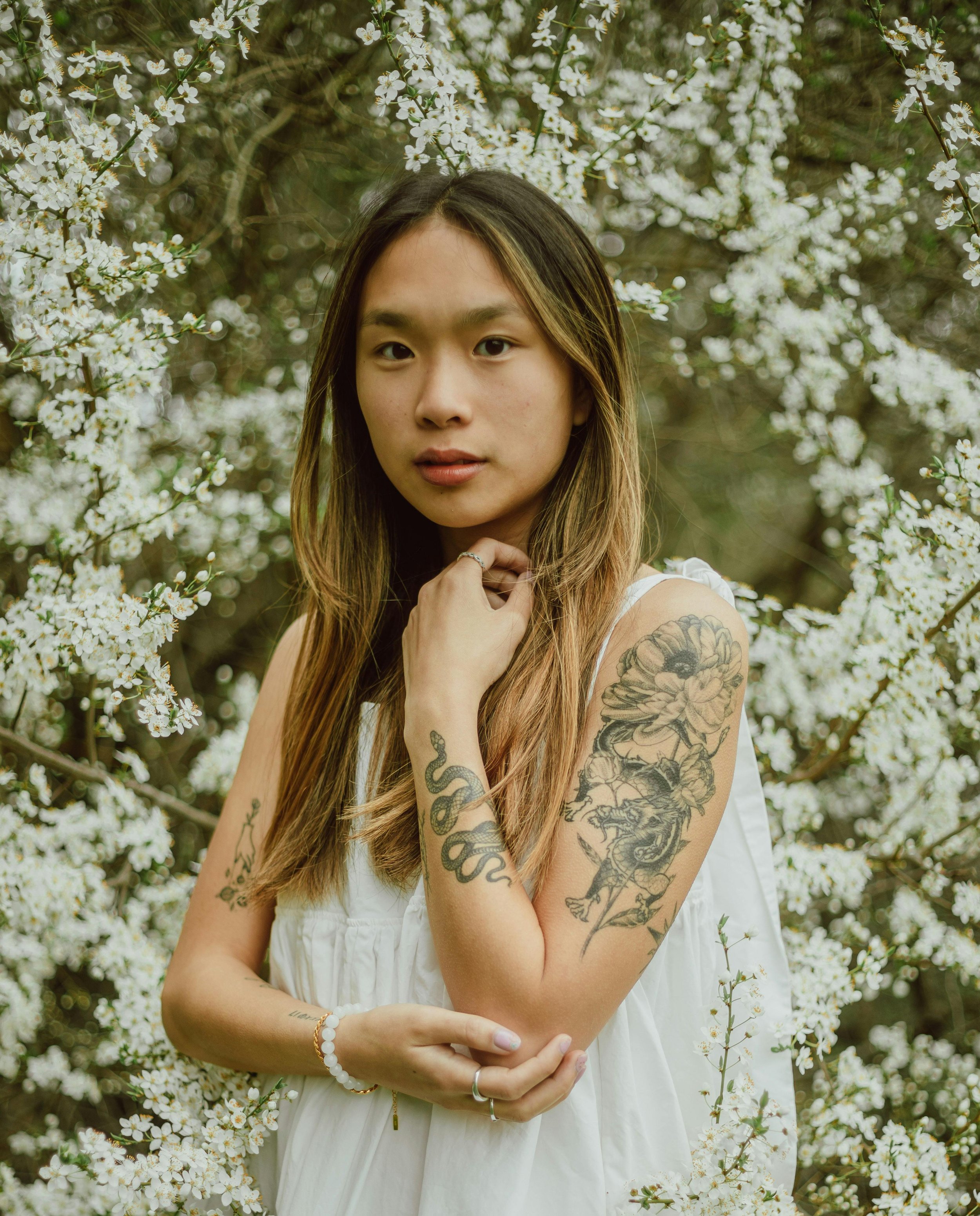 A young woman with long brown hair and tattoos on her arm is standing among white flowering trees. She is wearing a white dress and jewelry, and looking at the camera.