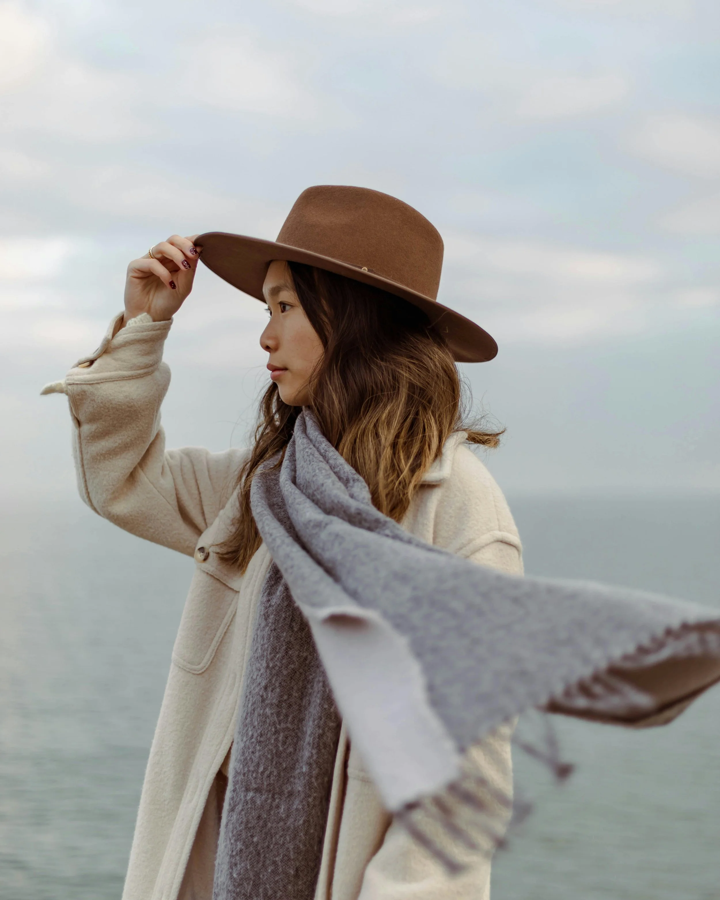 A young woman with long brown hair wearing a brown wide-brimmed hat, a light beige coat, and a gray scarf, standing outdoors near a body of water with a cloudy sky in the background.