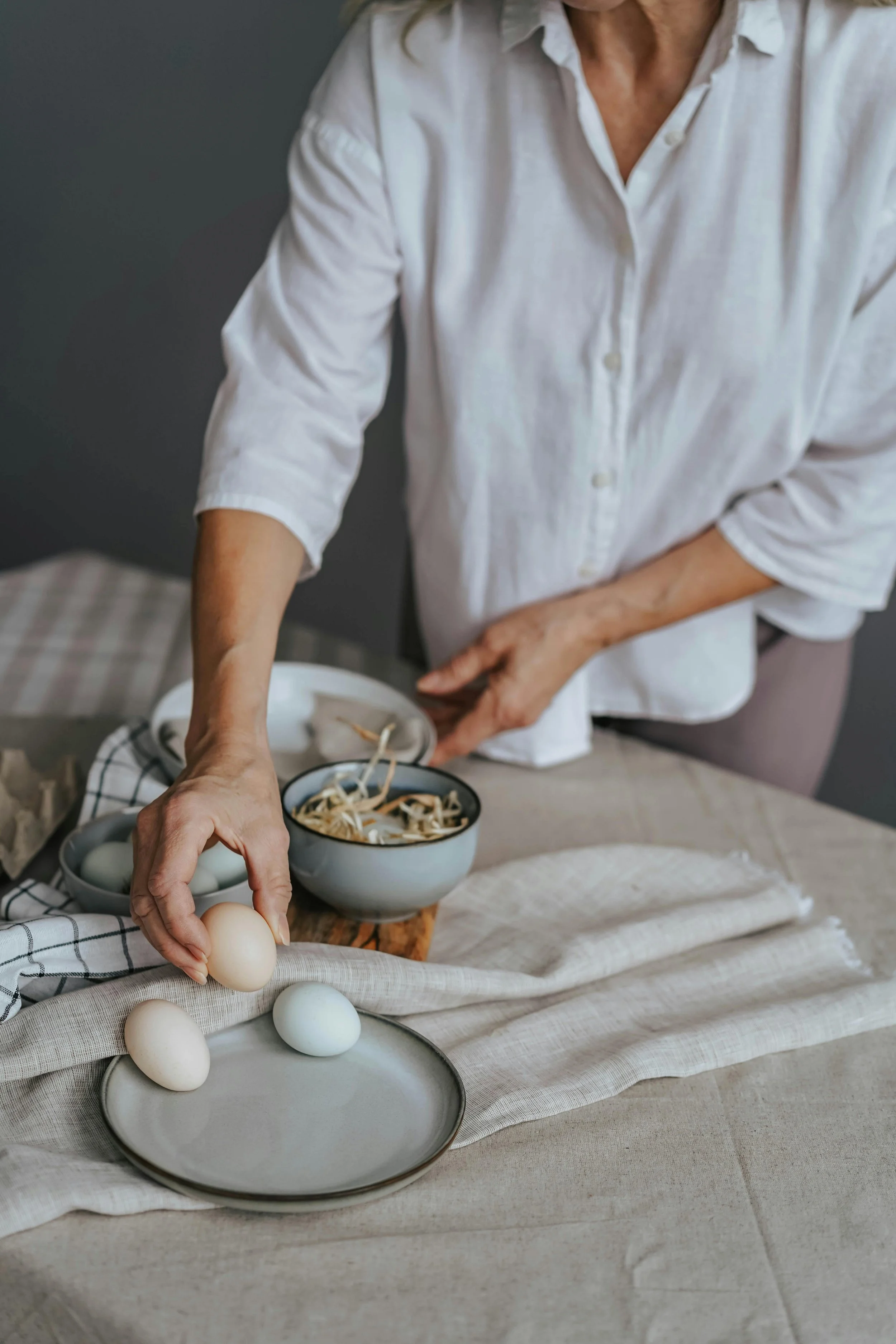 Person in a white shirt placing eggs on a table with a bowl of sliced sprouts and a bowl of eggs, on a beige tablecloth.