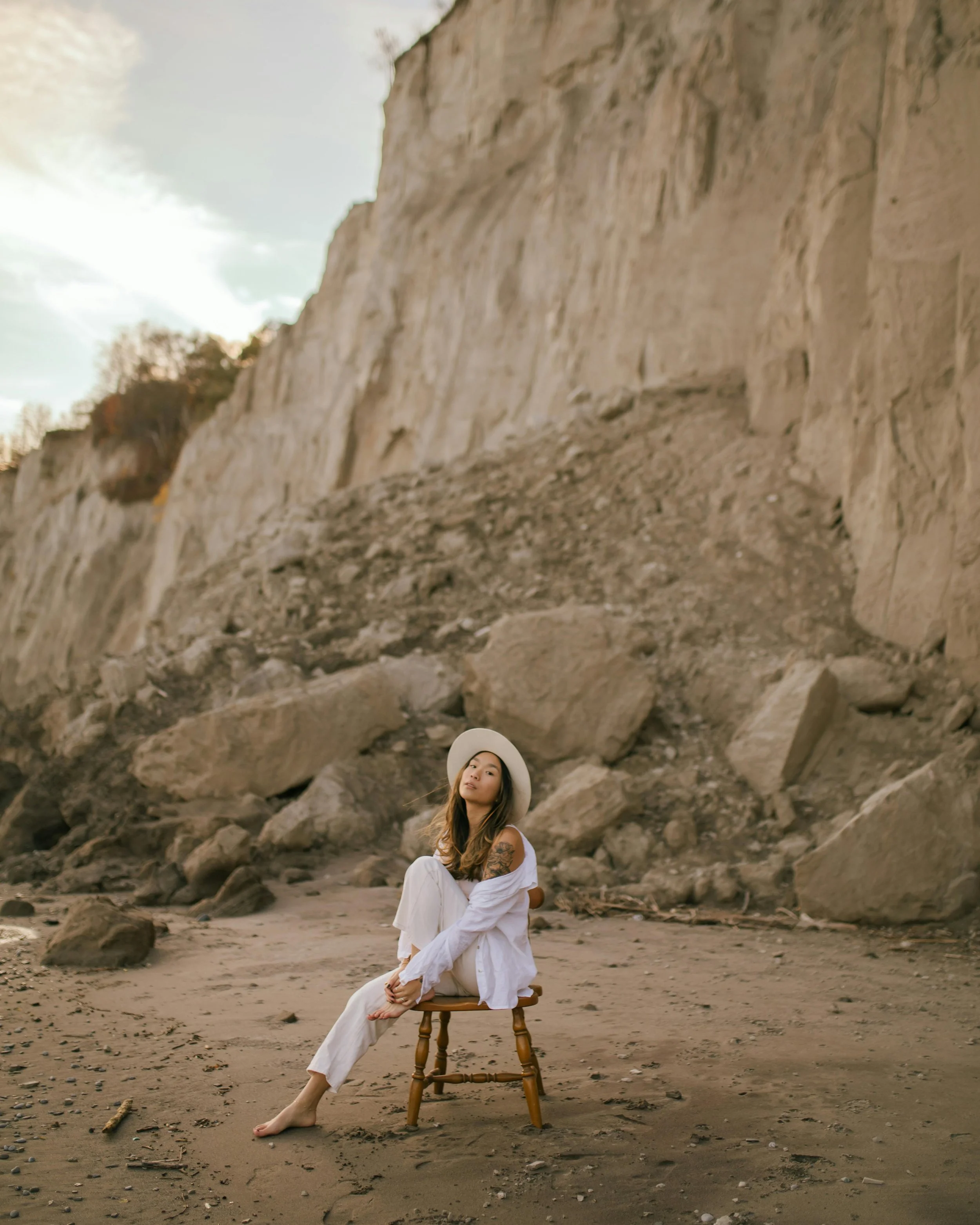 A woman with long hair and tattoos sitting on a small wooden stool on a beach, wearing a white hat and clothing, with a large rock formation and cloudy sky in the background.