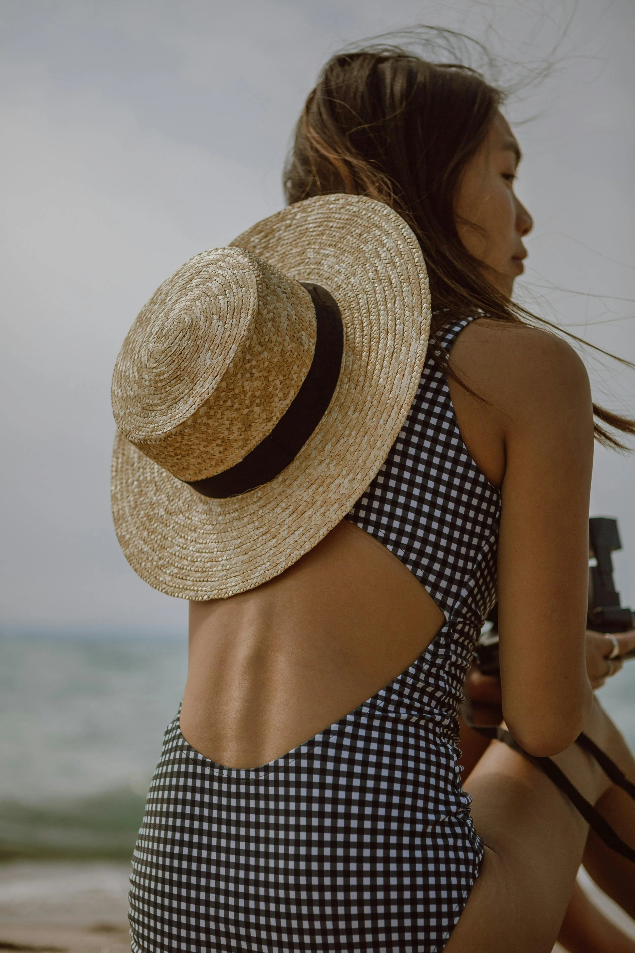 Young woman with long hair wearing a gingham swimsuit and holding a straw hat, standing on the beach with the ocean in the background.
