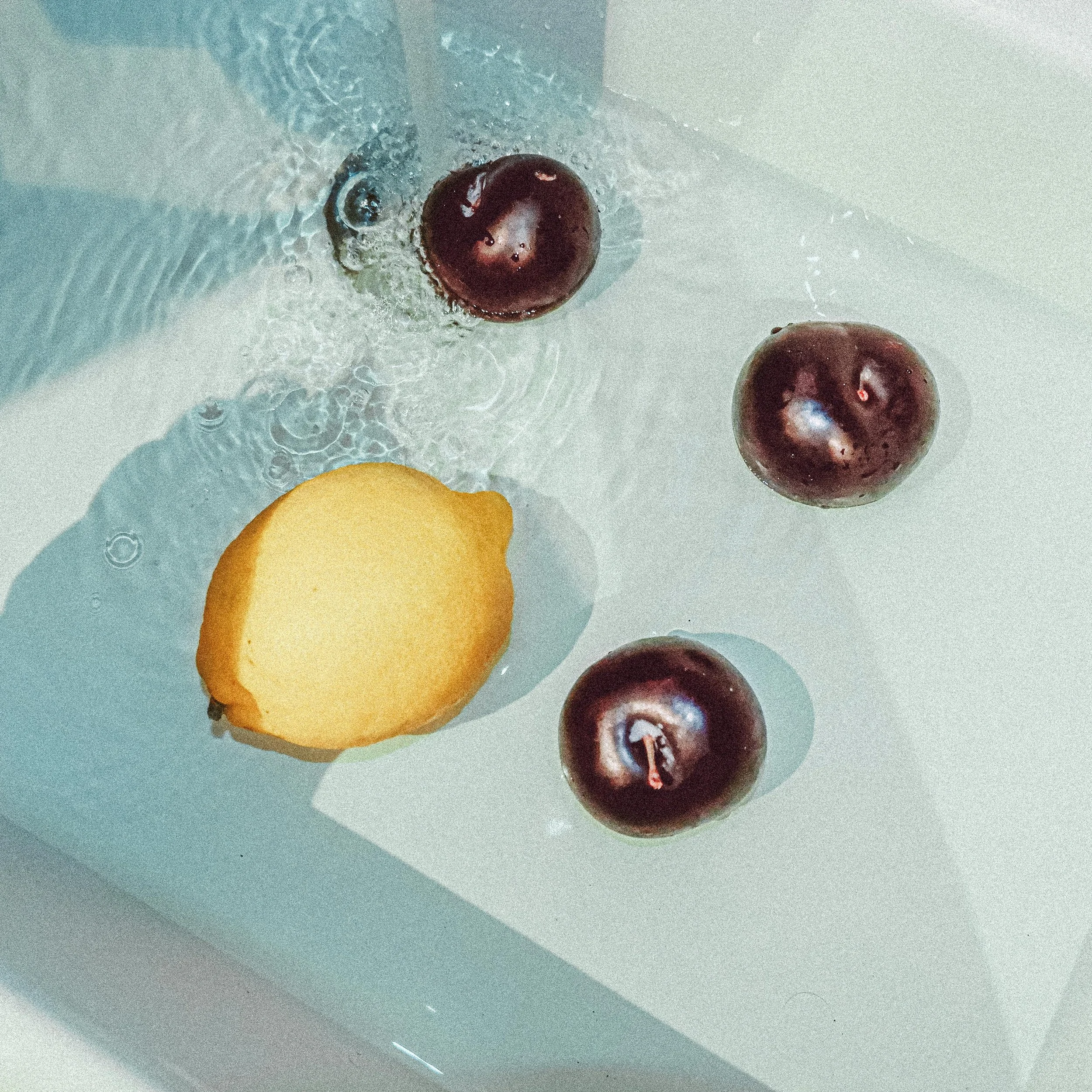 Close-up of two plums and one lemon in a sink with water running, creating ripples around the fruit.
