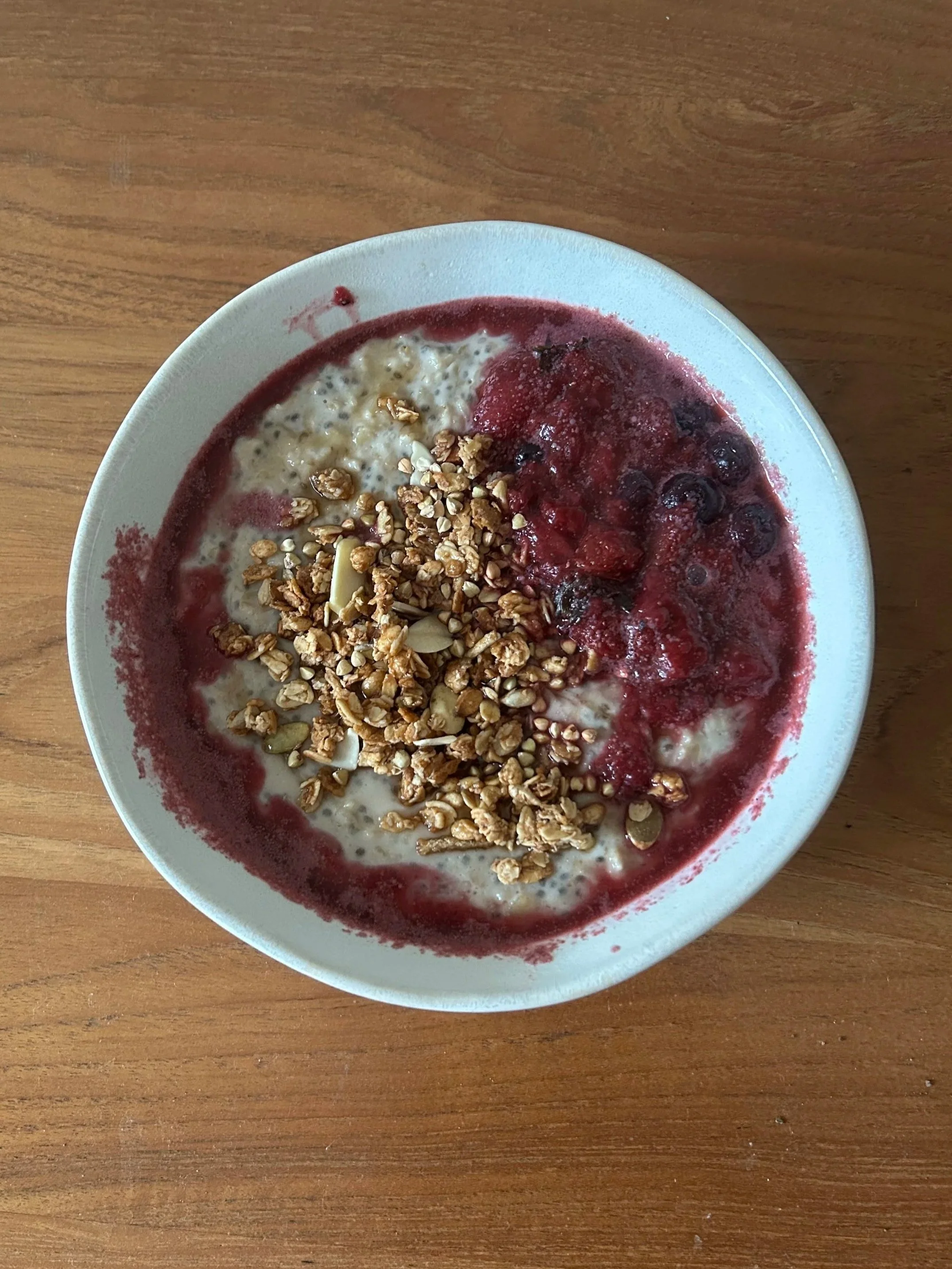 A bowl of yogurt topped with granola, nuts, and a berry sauce, placed on a wooden surface.
