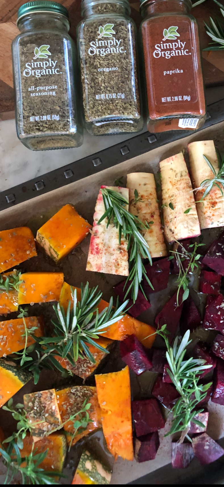 Three jars of organic spices labeled all-purpose seasoning, oregano, and paprika, with assorted roasted vegetables garnished with herbs on a tray.